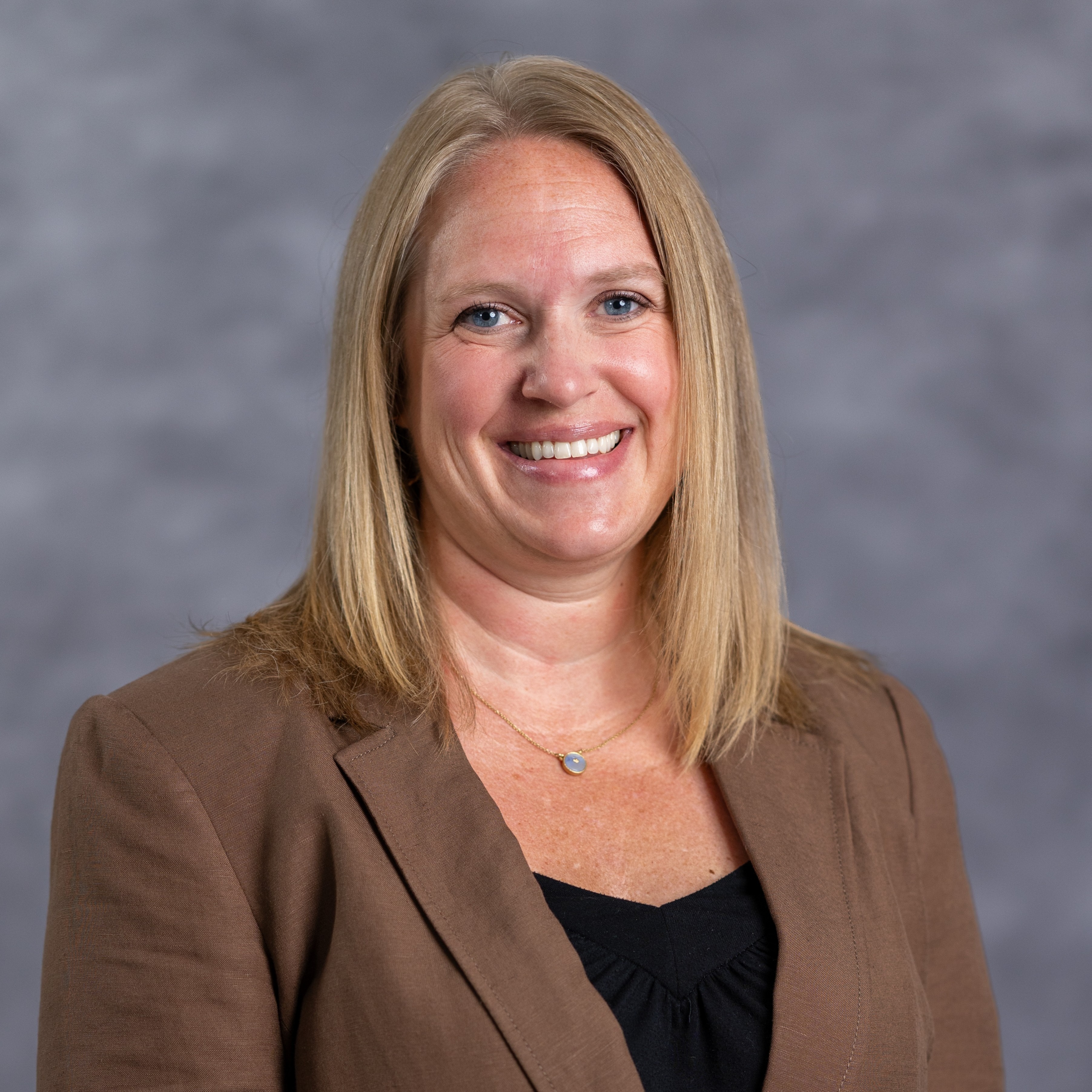 Woman with shoulder-length blond hair, smiling, wearing a brown blazer over a black top.