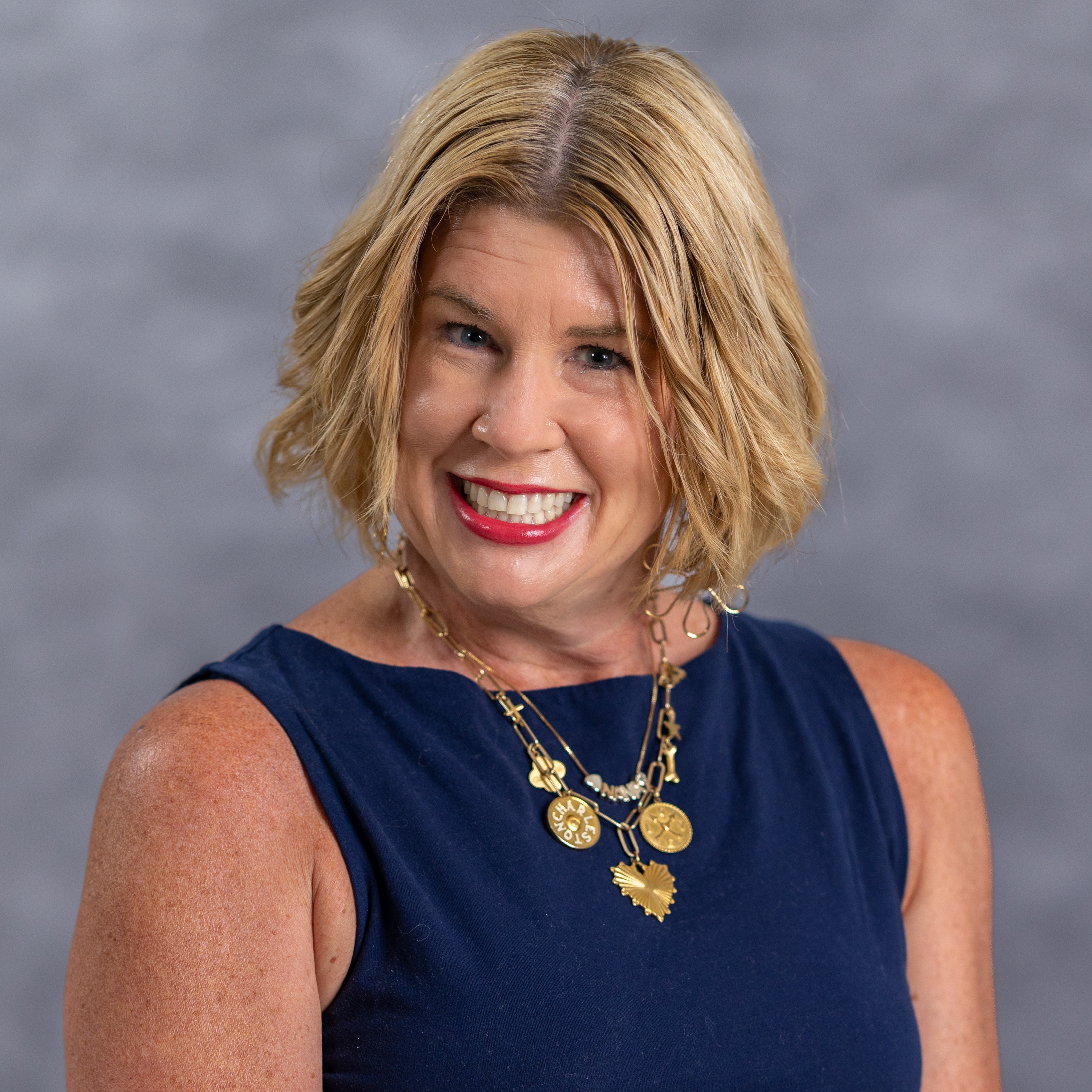 A woman with short blonde hair, wearing a navy sleeveless top, smiles at the camera. She is adorned with a gold necklace featuring heart-shaped pendants.