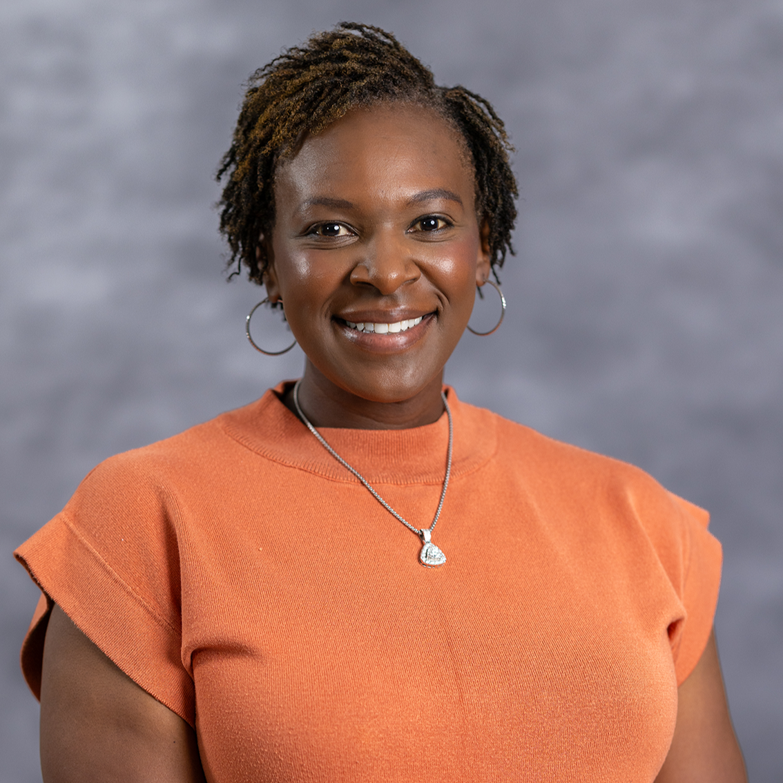 Woman with braided hair in an orange top, smiling at the camera. She wears a necklace with a heart pendant.