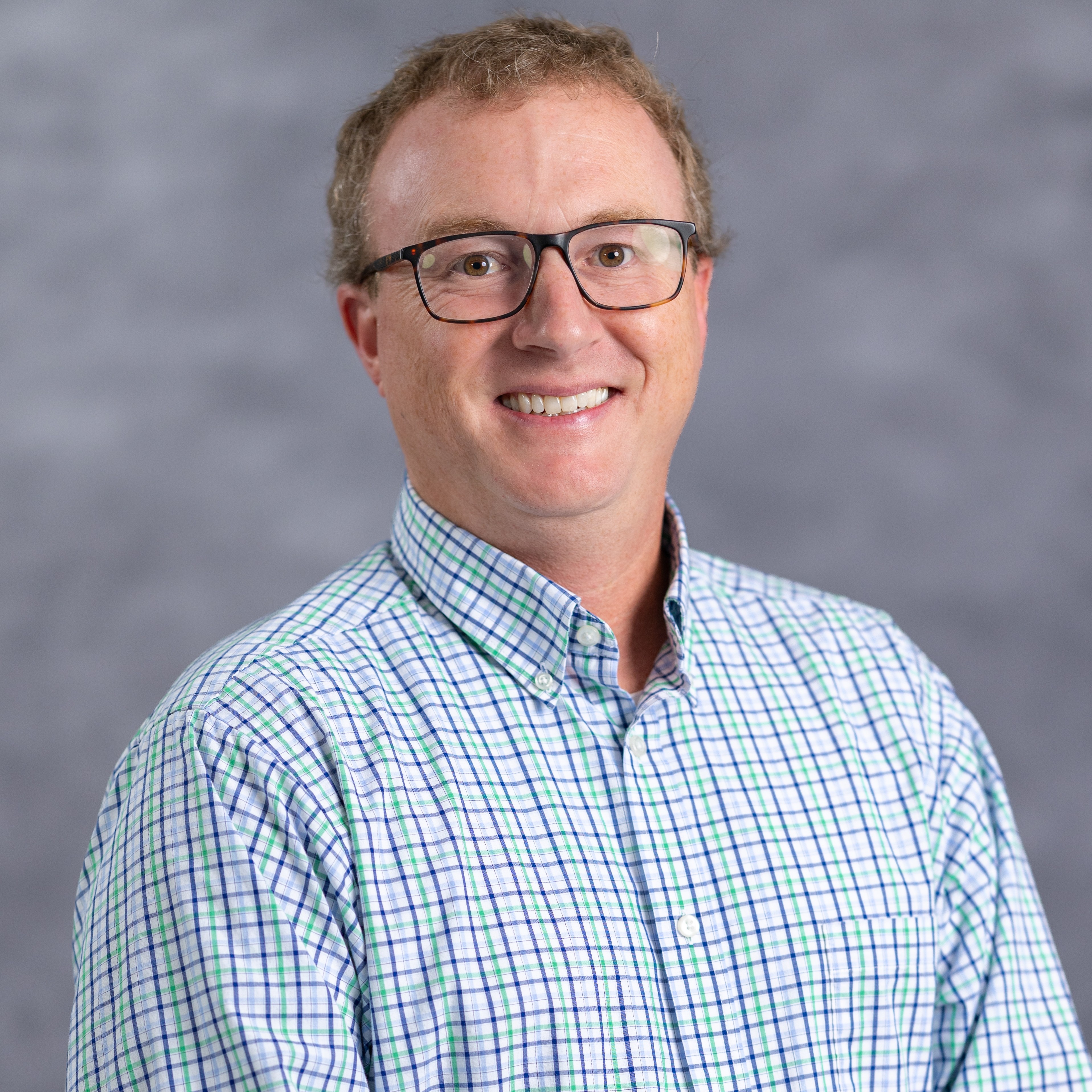 A smiling man in a blue and white checked shirt with glasses stands against a gray background.