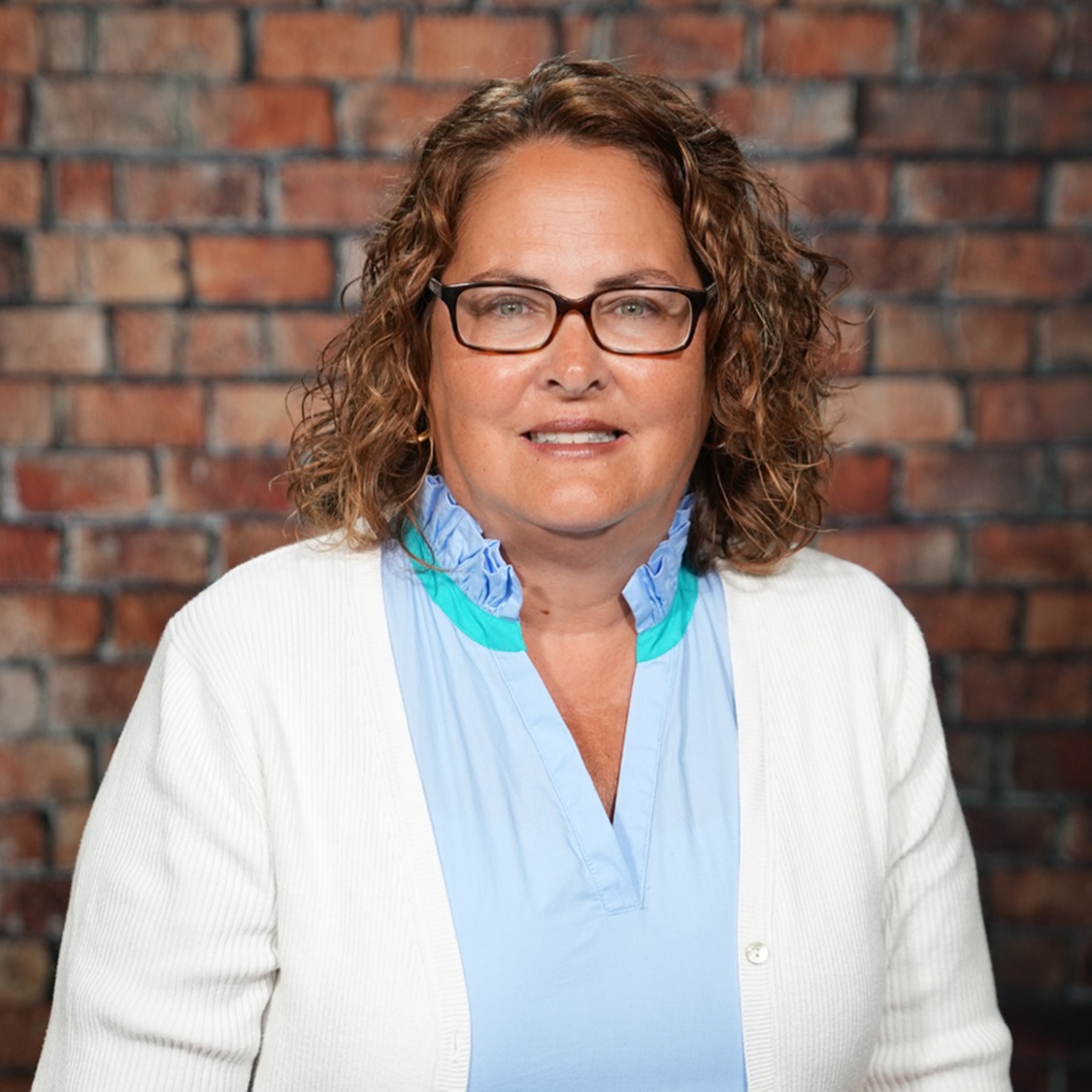 Woman with curly hair wearing a white cardigan and blue collared shirt stands before a brick wall.