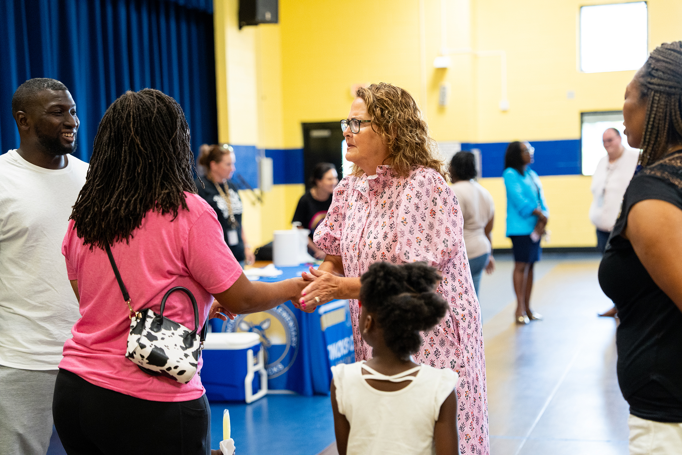 Multiple adults and children interact in an indoor space. One woman shakes hands with another woman.