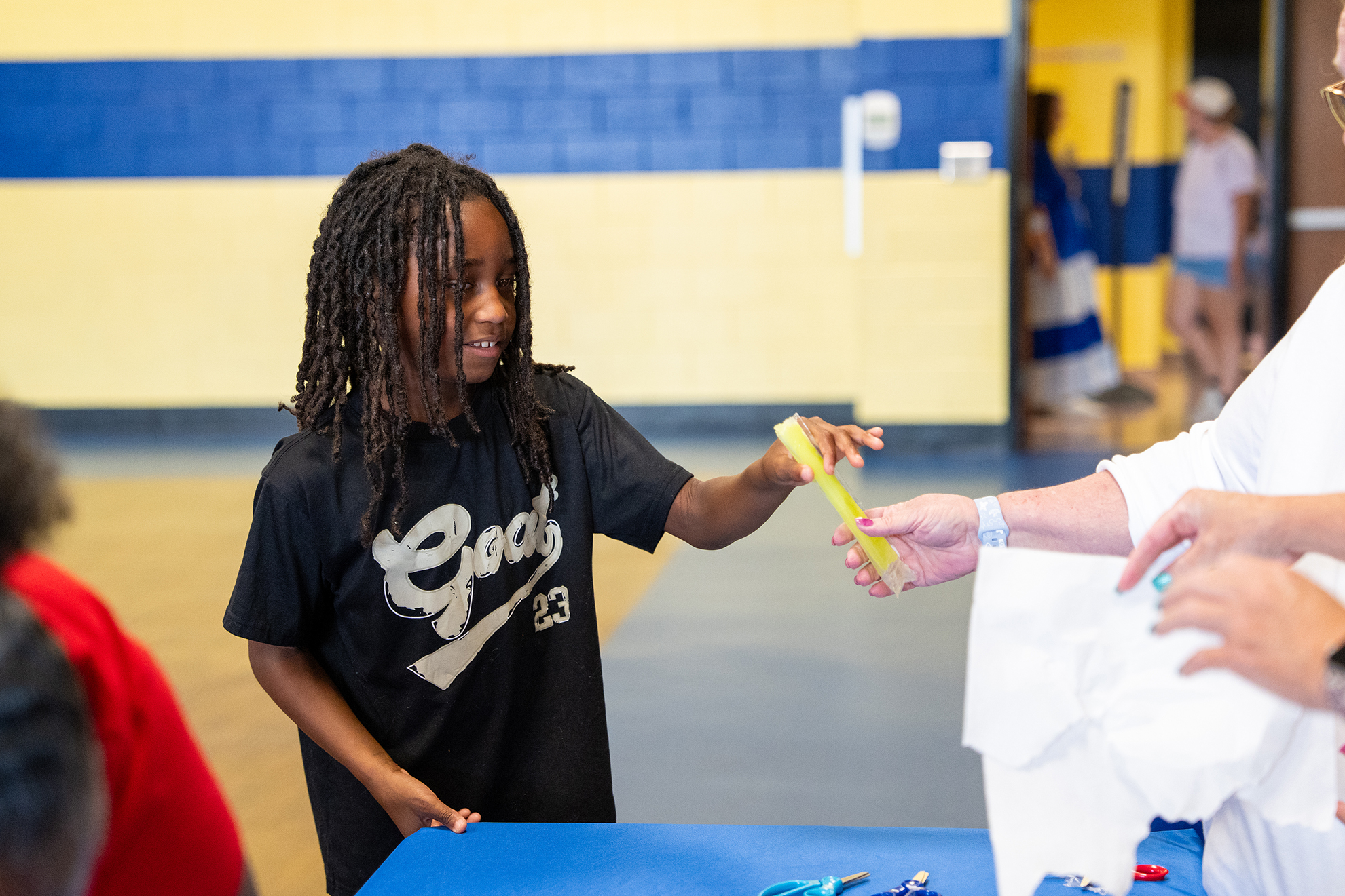 A child with braided hair holds a yellow object, smiling, as an adult offers a white cloth.