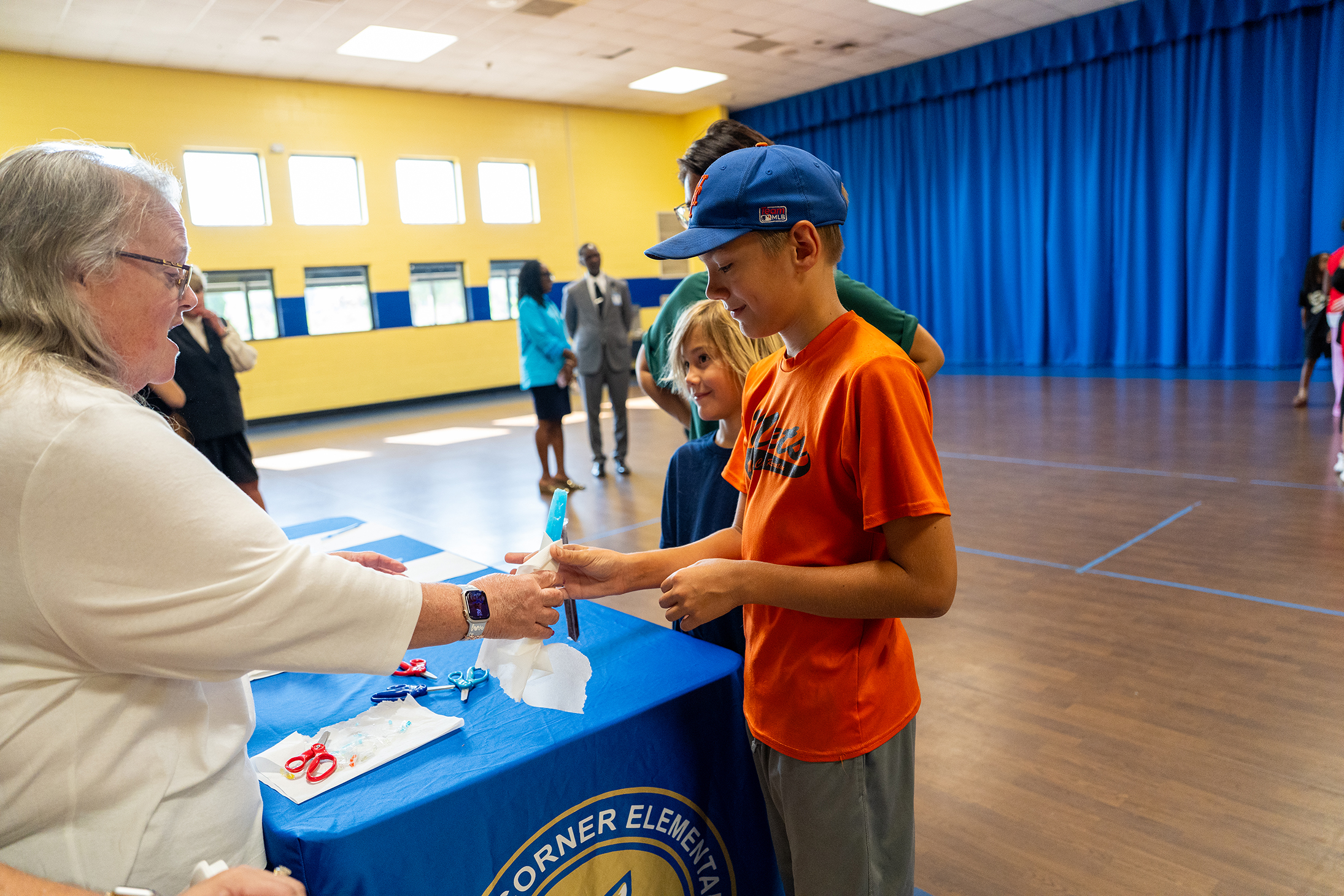 A woman hands a boy a clear plastic item at a table in a gymnasium. Other people are present in the background.