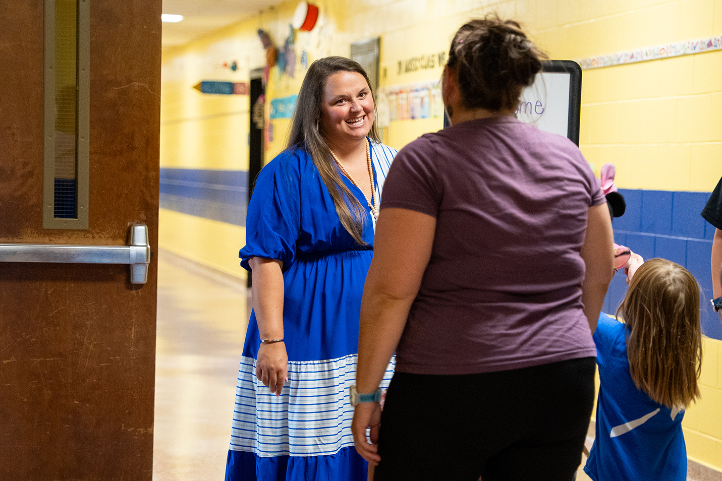 A woman in a blue dress converses with another woman in a purple shirt in a hallway.