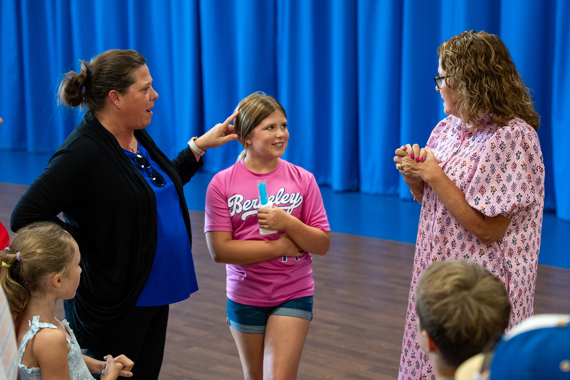 Several adults and children are gathered indoors. A woman holds a young girl's hair while another woman looks on.