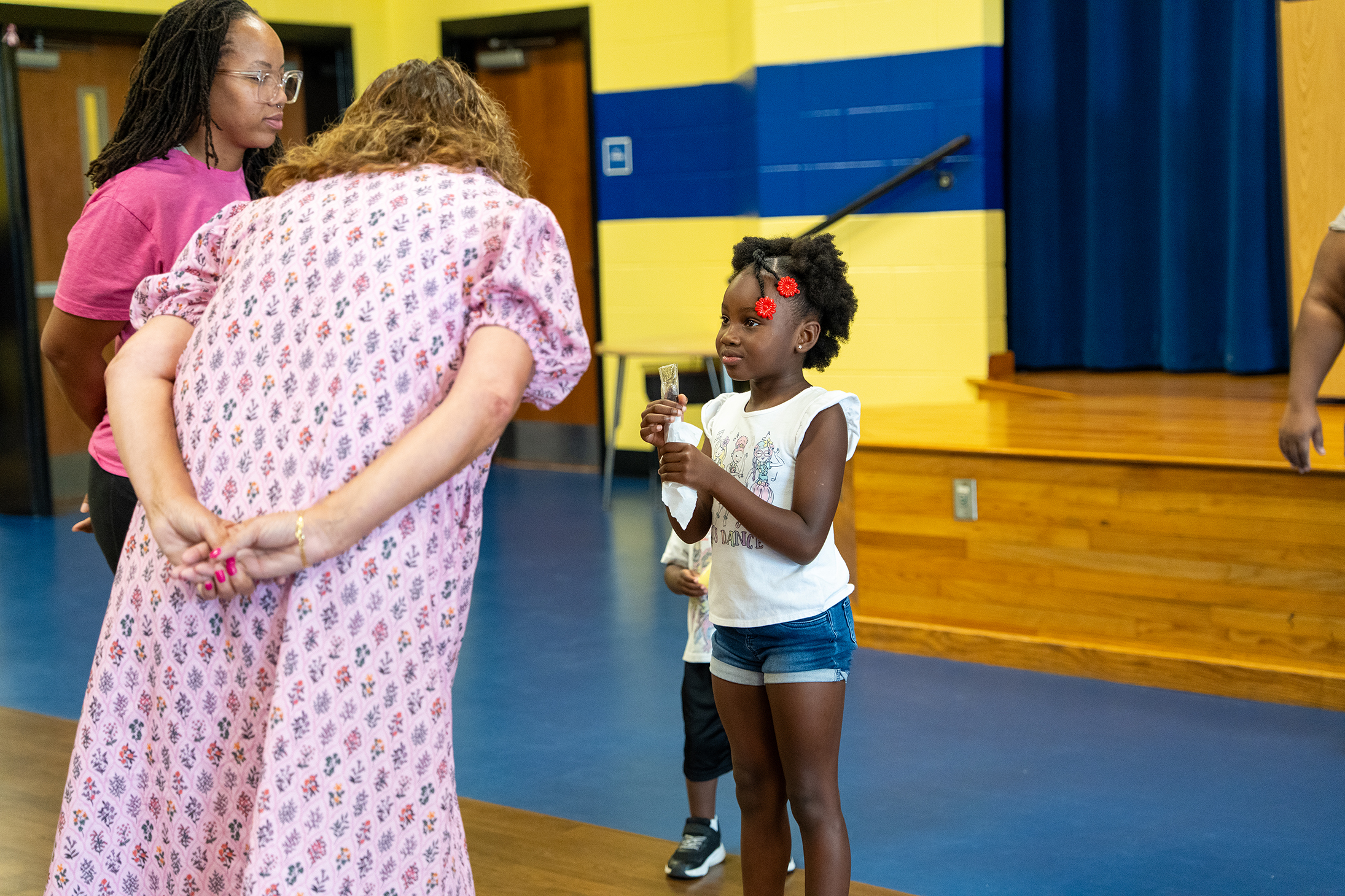 A girl in a white shirt and shorts holds a microphone, standing on a stage with a wooden floor. Behind her, a woman in pink holds her back.