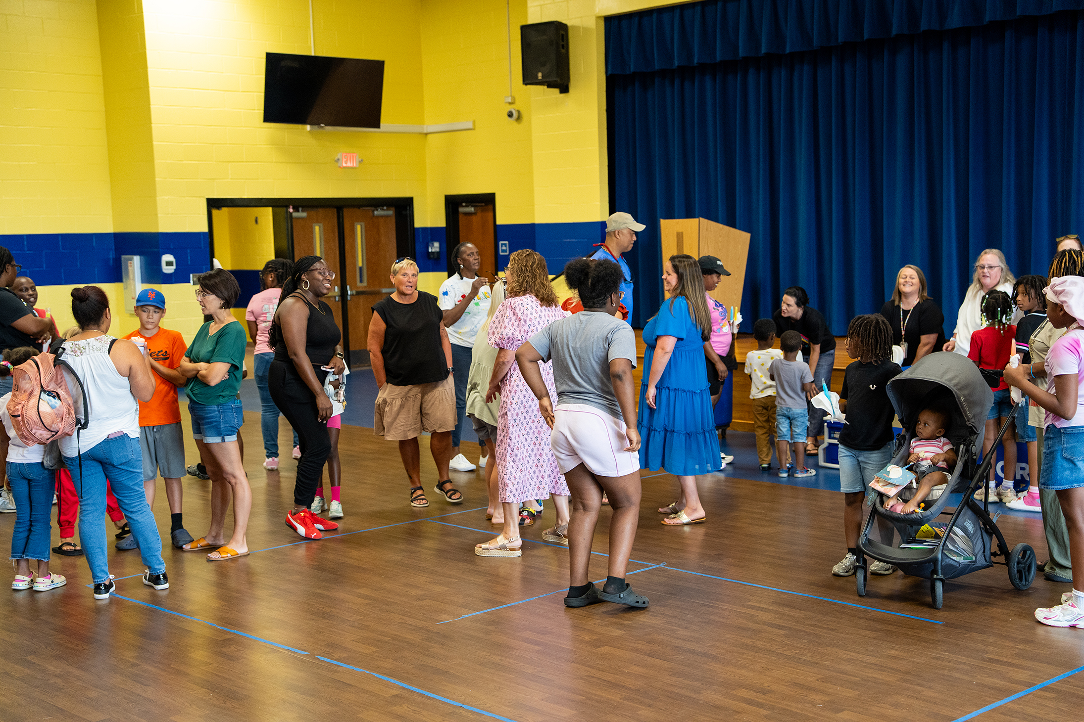 A hall with a wooden floor and yellow walls. People stand and converse. A stroller is nearby.