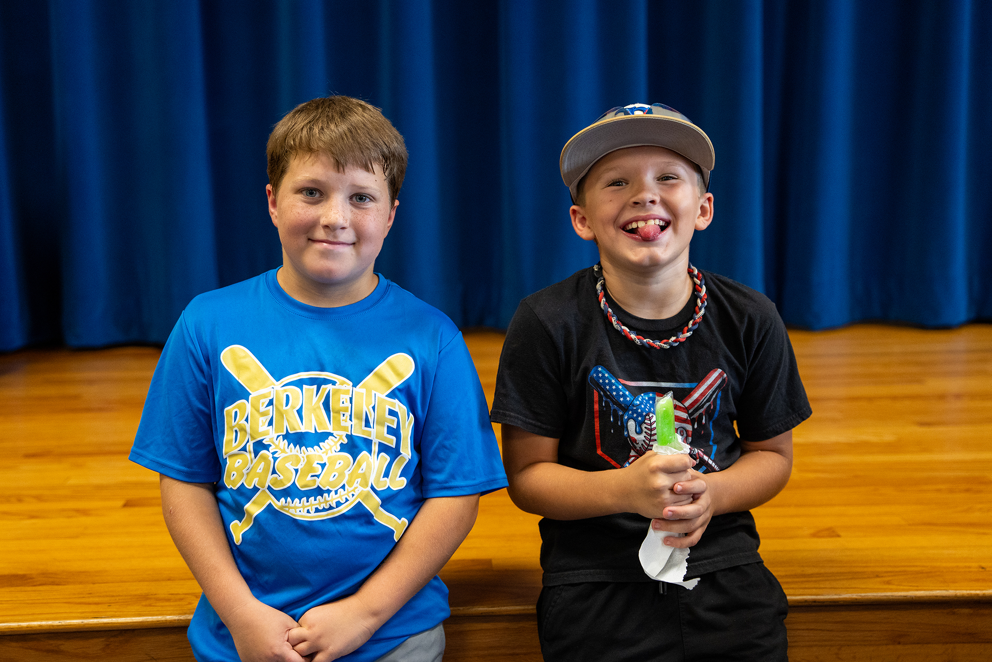 Two young boys, one in a blue shirt and the other in black, sit next to each other on a stage.