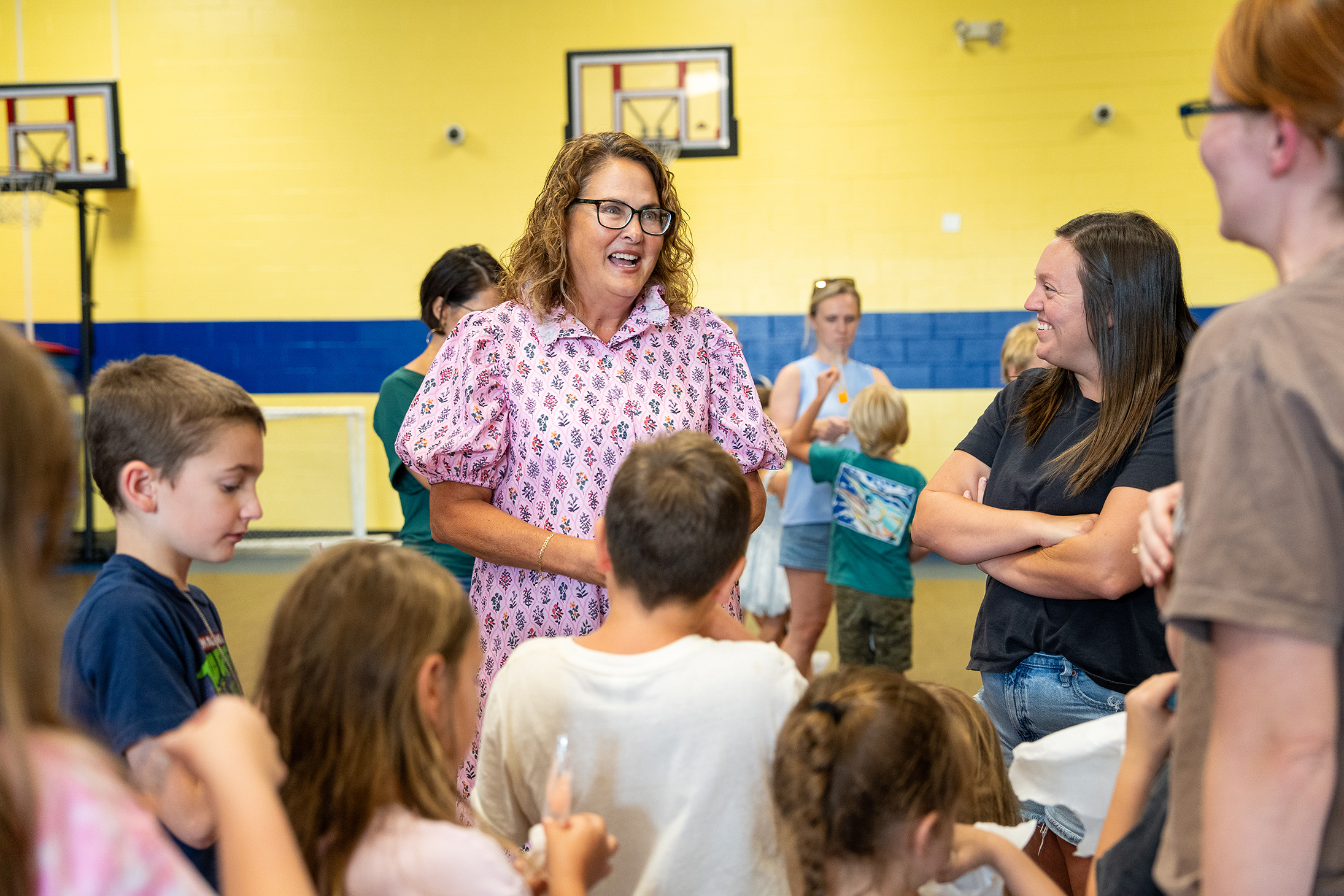 People gather in a gym, including adults and children. A woman in pink stands with others.