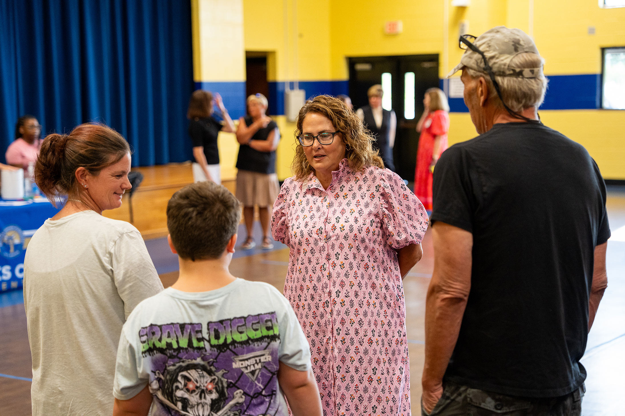 A group of adults and children are conversing in a room with yellow and blue walls, a blue curtain, and a wooden floor.