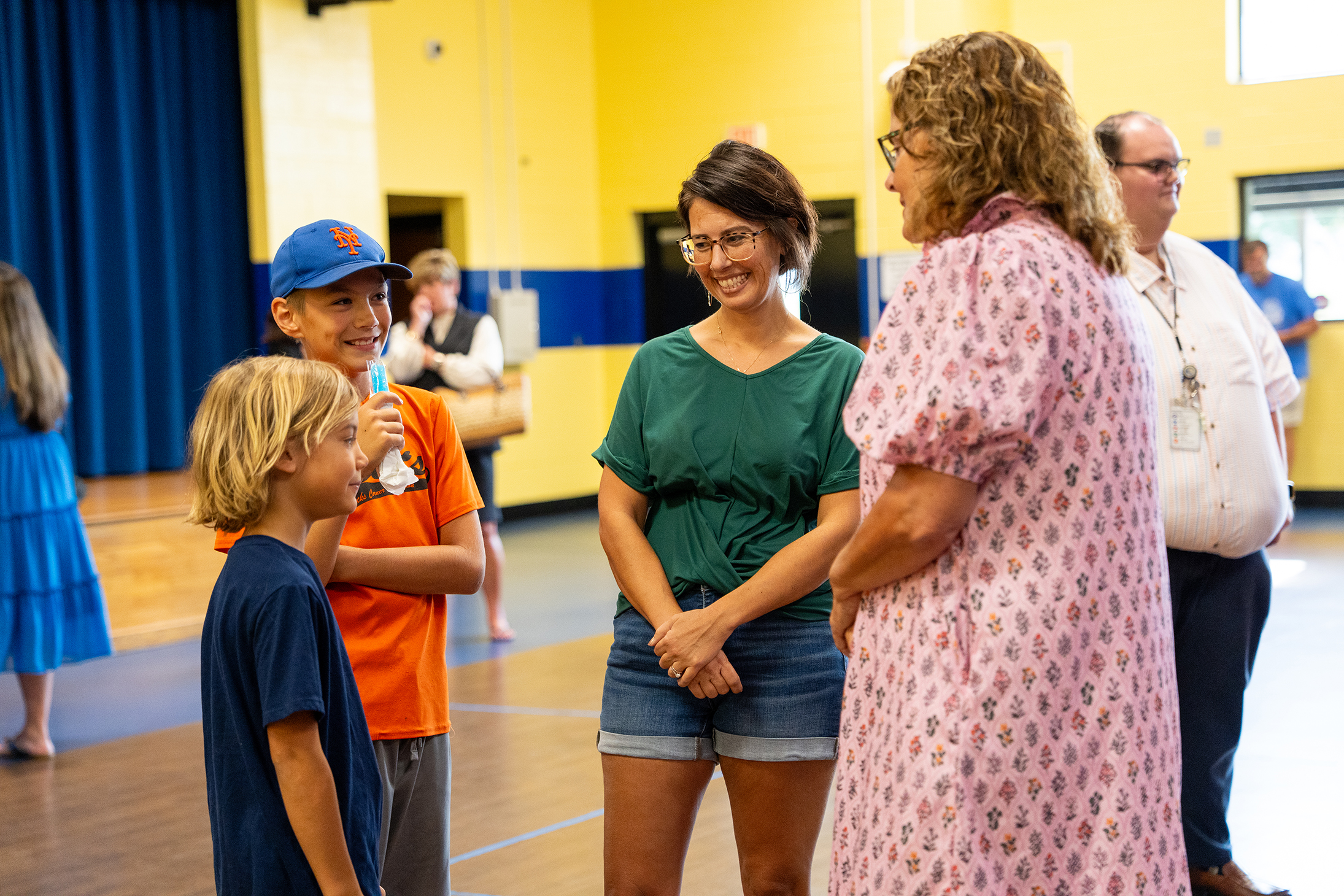 Five people converse in a room with yellow and blue walls. A woman in a pink dress and a boy wear glasses.