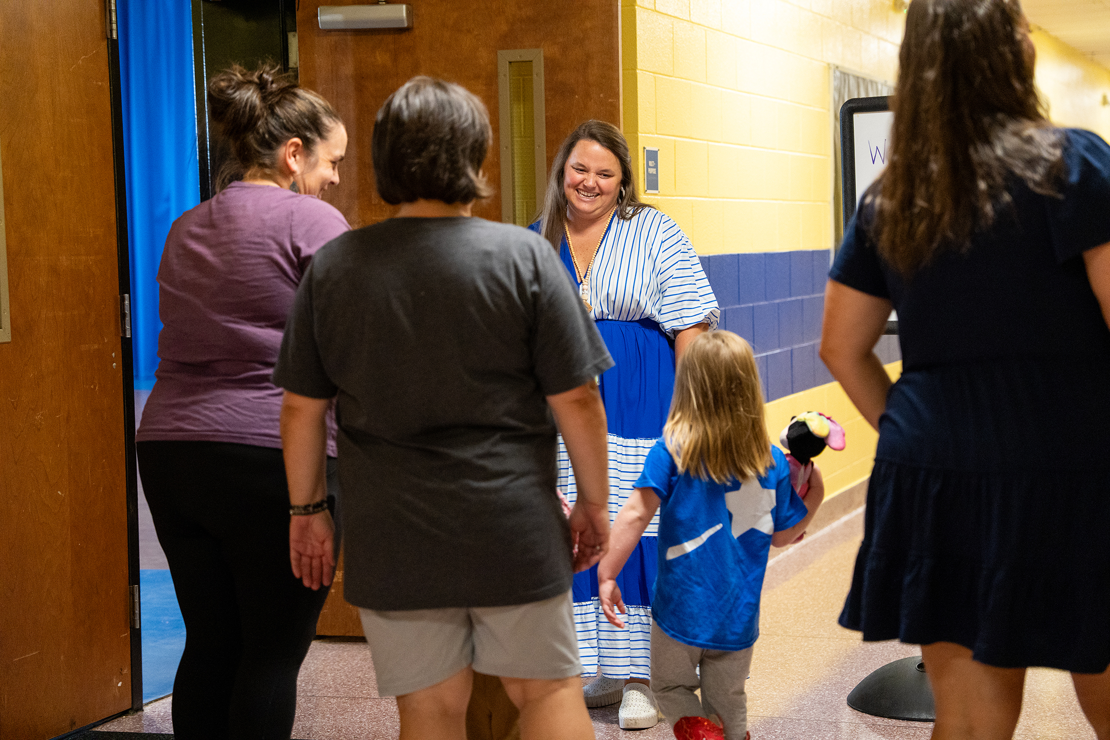 A group of people stand in a hallway; a woman in a blue dress leads a child.