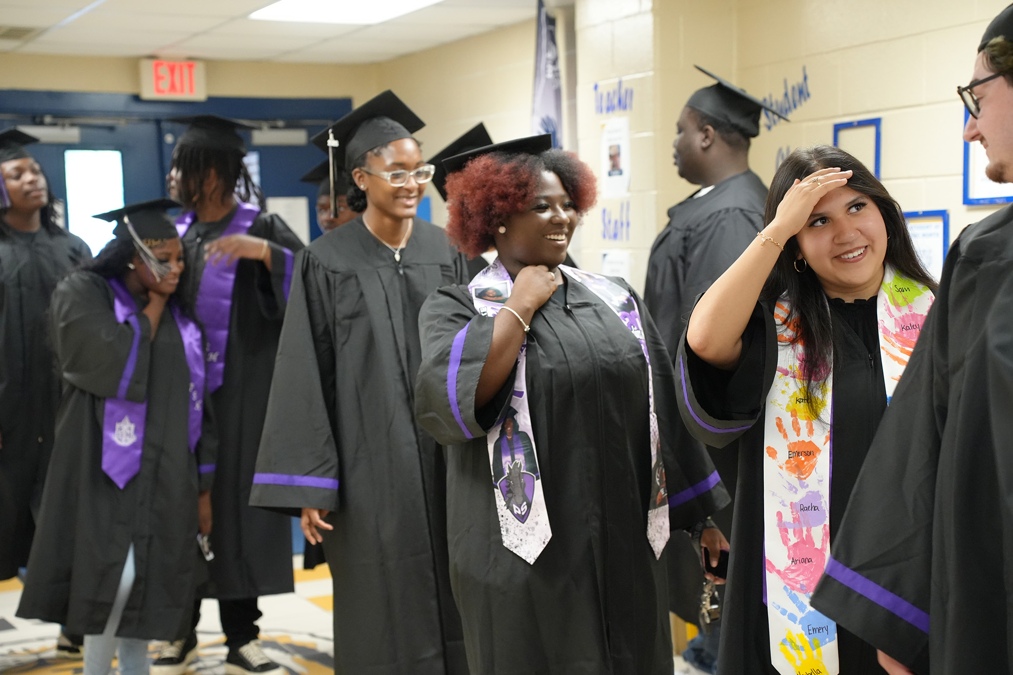 People in graduation gowns and caps stand in a line, with one woman adjusting her gown.