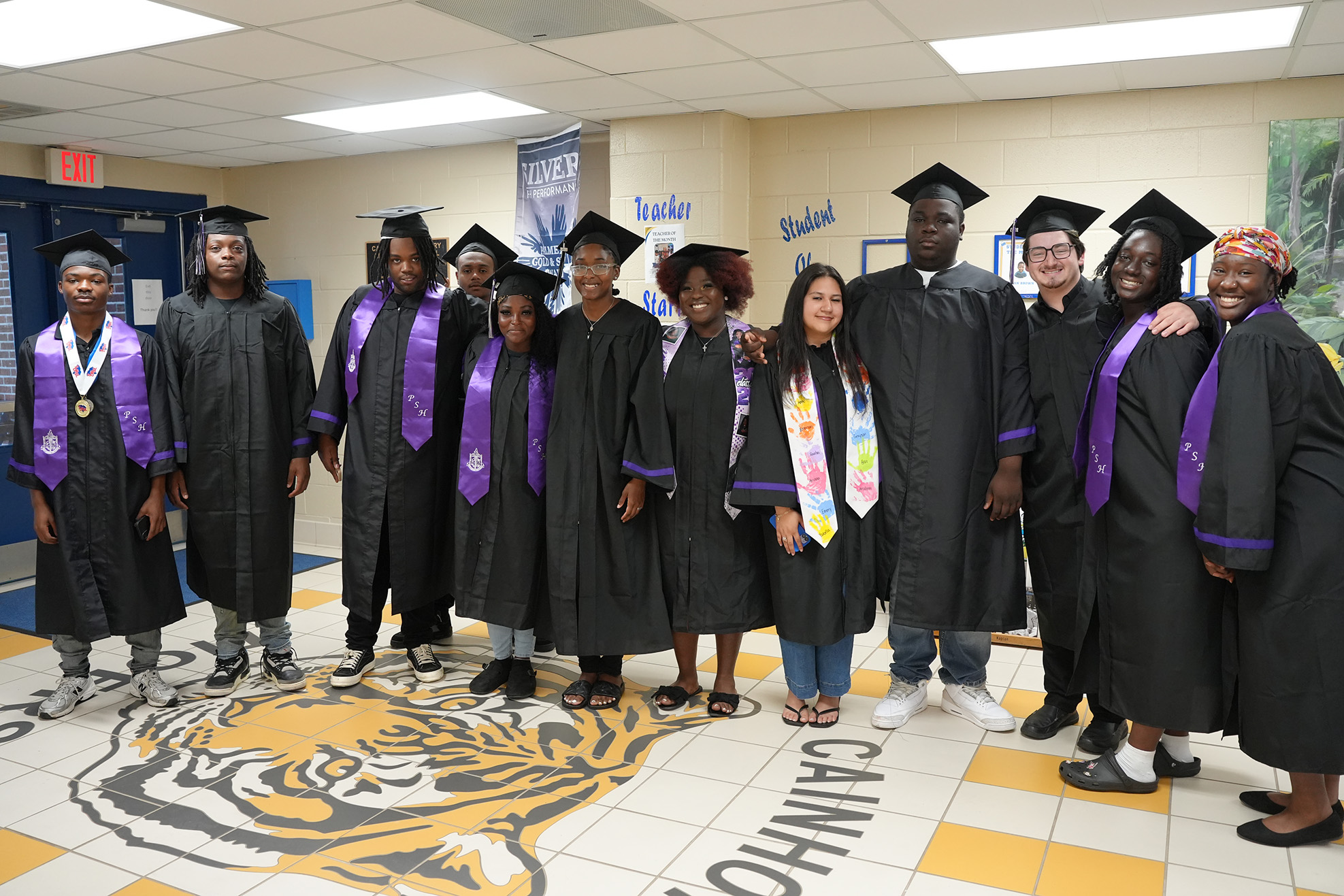 A group of people in graduation gowns and caps stand in a hallway. Some wear sashes. The floor has a tiger logo.