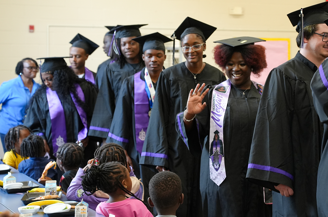 Several women in graduation gowns line up. Some wear medals and sashes. A few children sit at a table.