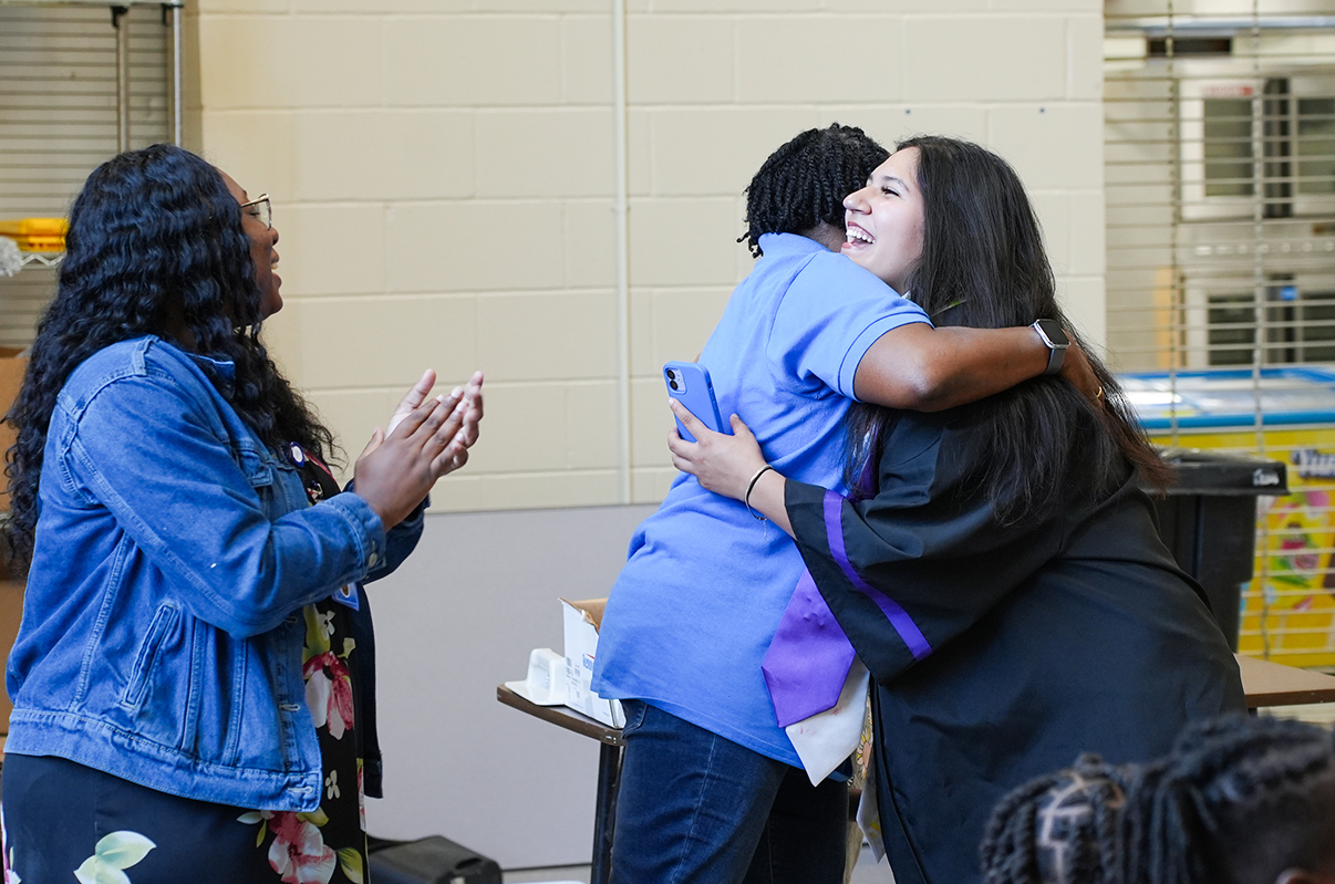 Three people stand in a room; one woman hugs another, both smiling, while a third woman claps. A table and trash bin are in the background.
