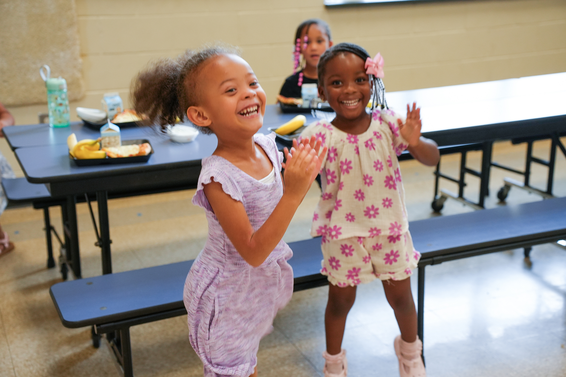 Two girls in a cafeteria. One is smiling and dancing. Behind them, a table has food, and another girl sits.