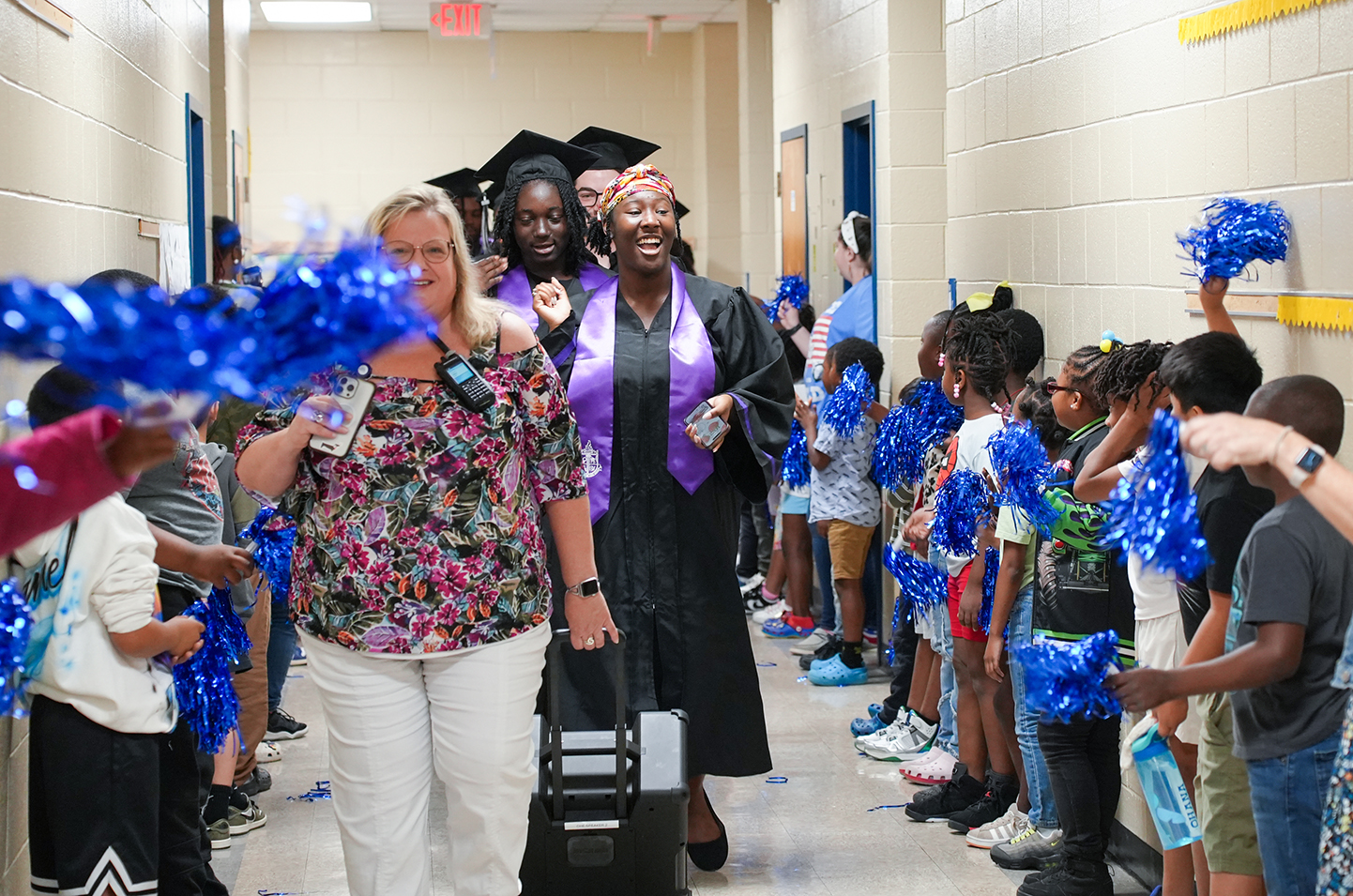 A group of people line a school corridor, with some holding blue pom-poms and cheering. A woman in a cap and gown walks with a suitcase.