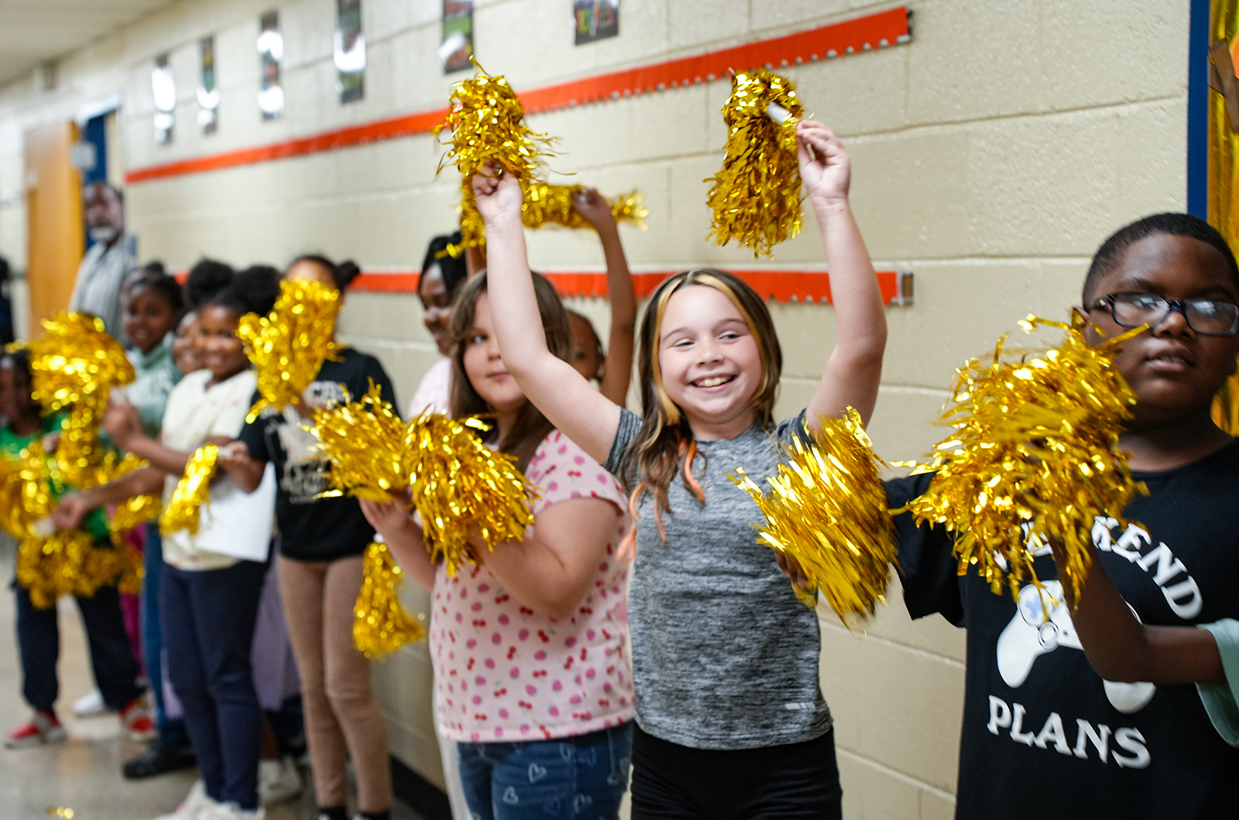 A group of young children holding gold pom-poms, smiling, with a hallway behind them.