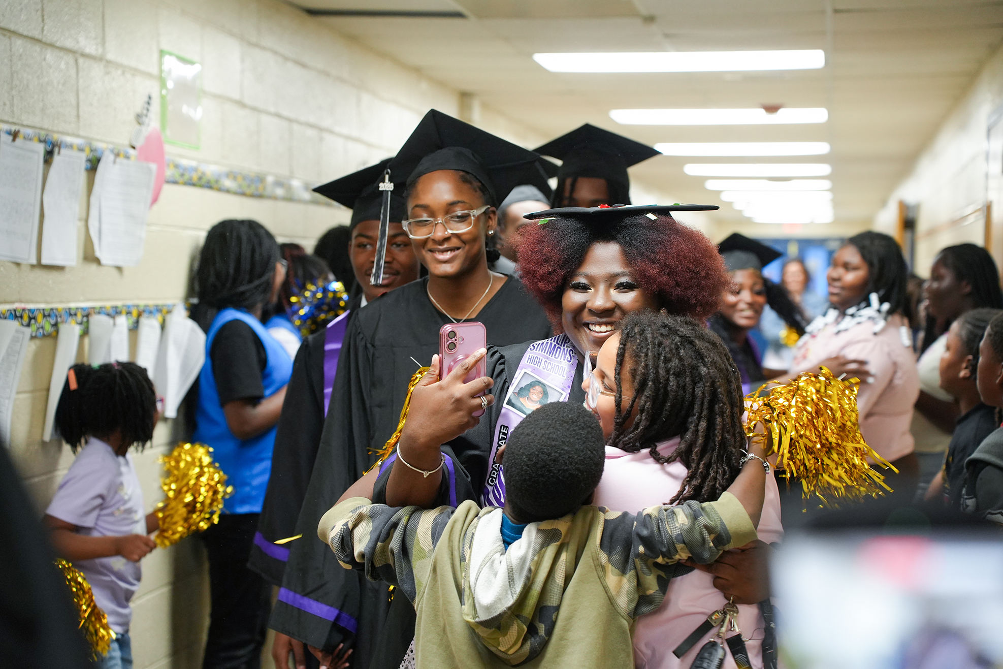 People gathered in a school hallway. One woman in a graduation gown smiles and holds a phone.