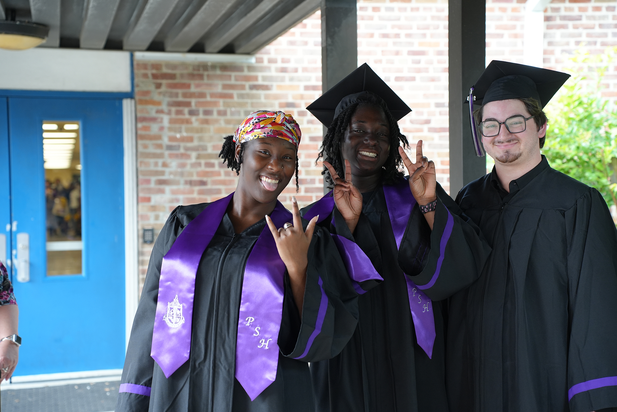 Three individuals in graduation attire, including caps, pose outside near a blue door. One person wears a purple sash.