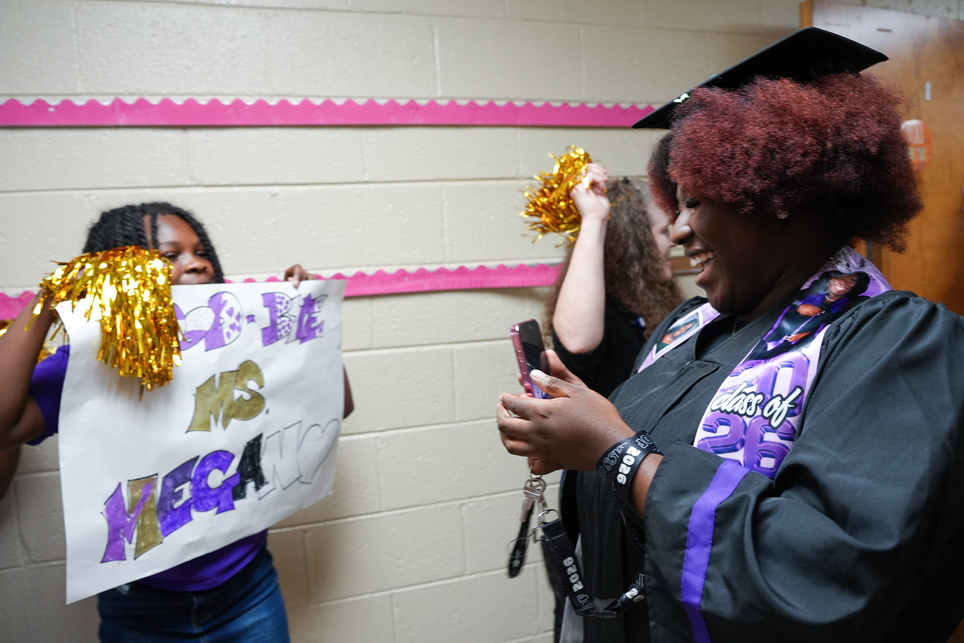 Two women in caps and gowns with a banner that says "WELCOME MS NECA" in front of a pink and white border.