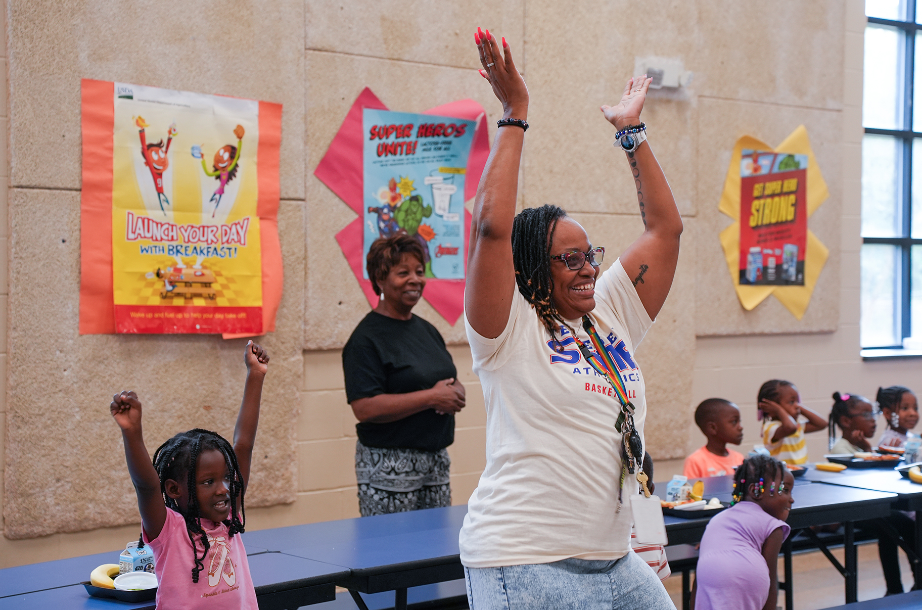 A woman and a young girl raise their arms inside a classroom. Several children sit at tables in the background.