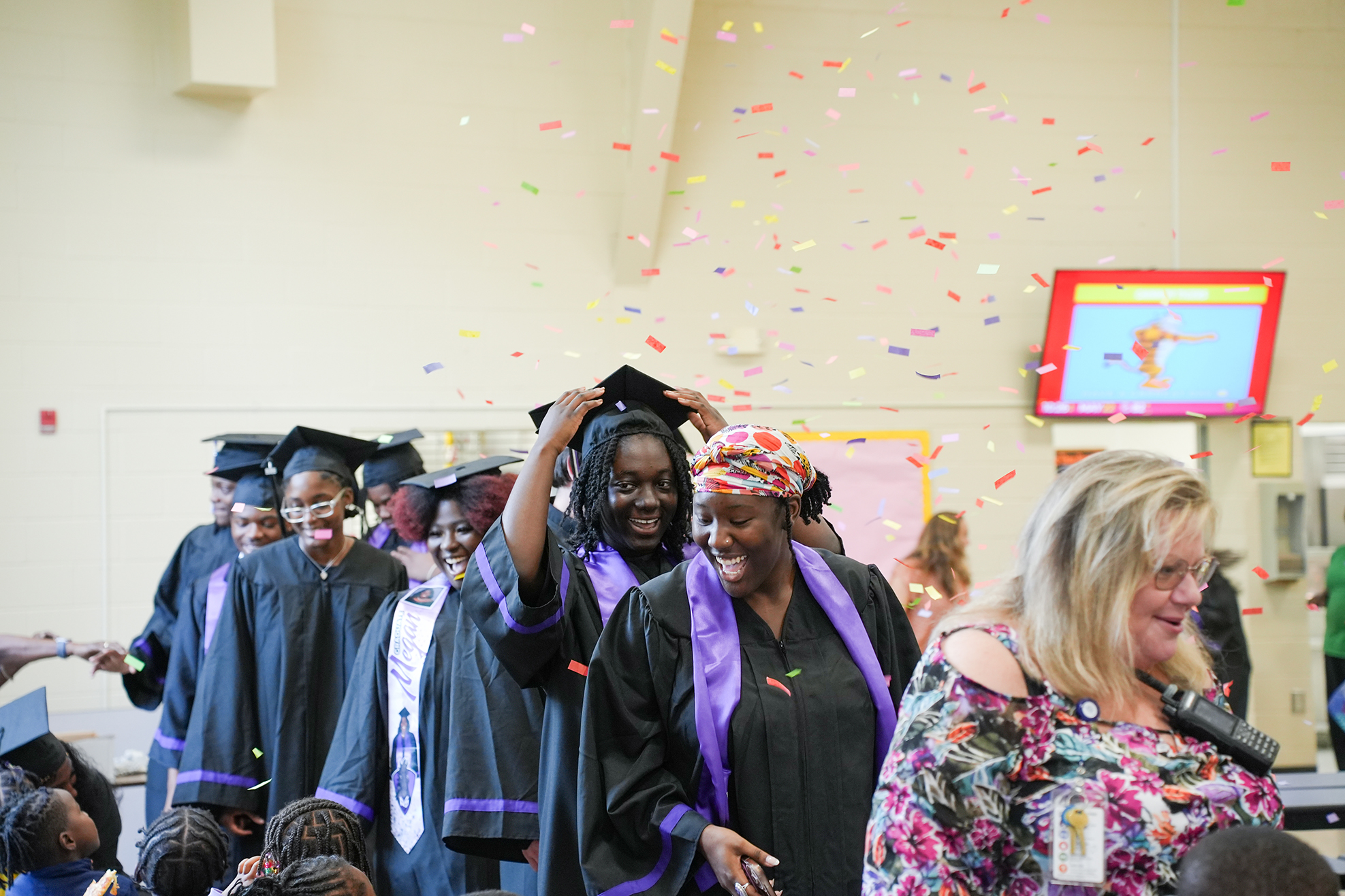 Graduates in caps and gowns celebrate in a room with confetti. Some wear head coverings.