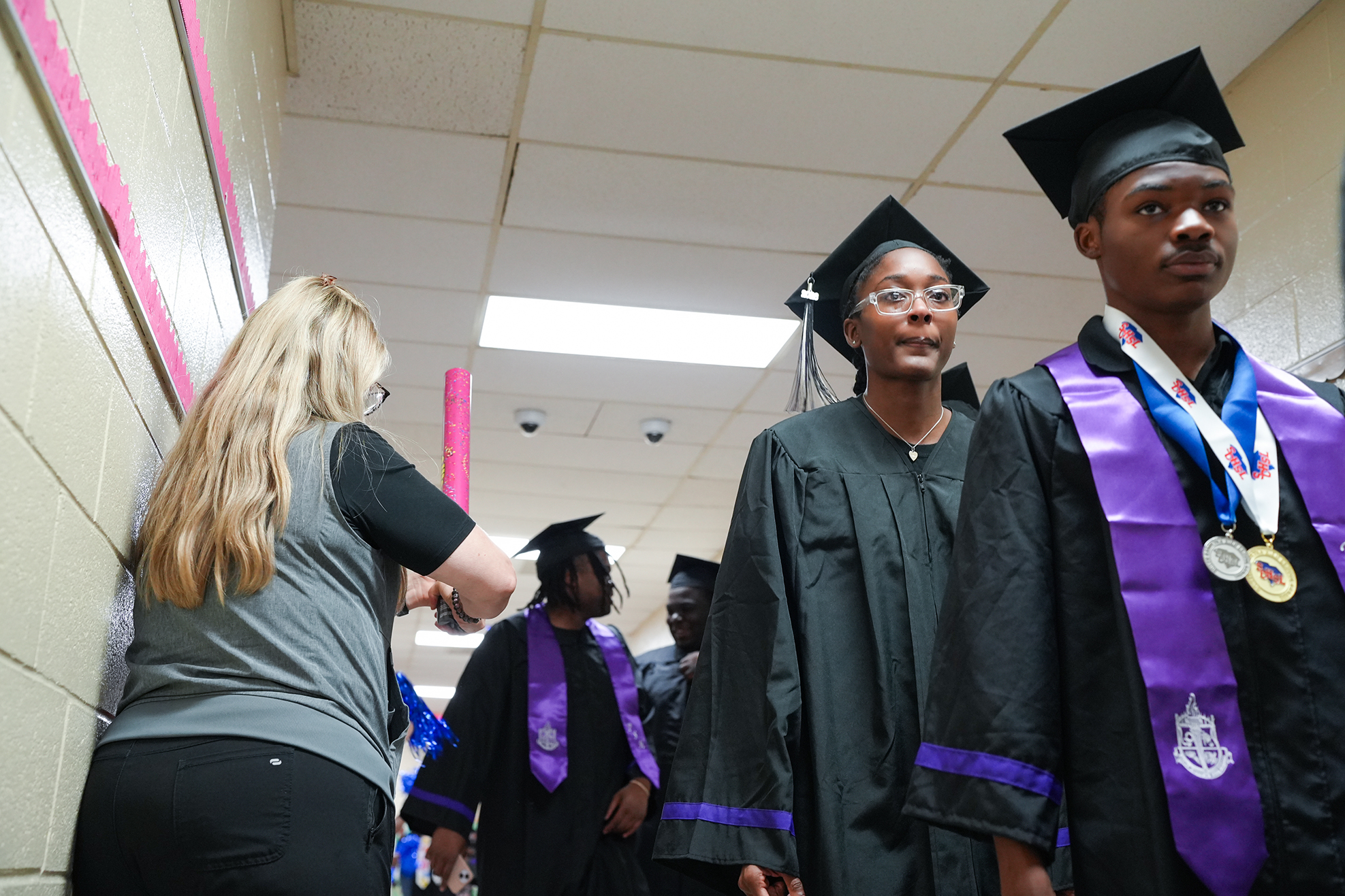 Graduates wearing caps and gowns walk down a hallway. A woman in a gray shirt looks at them.