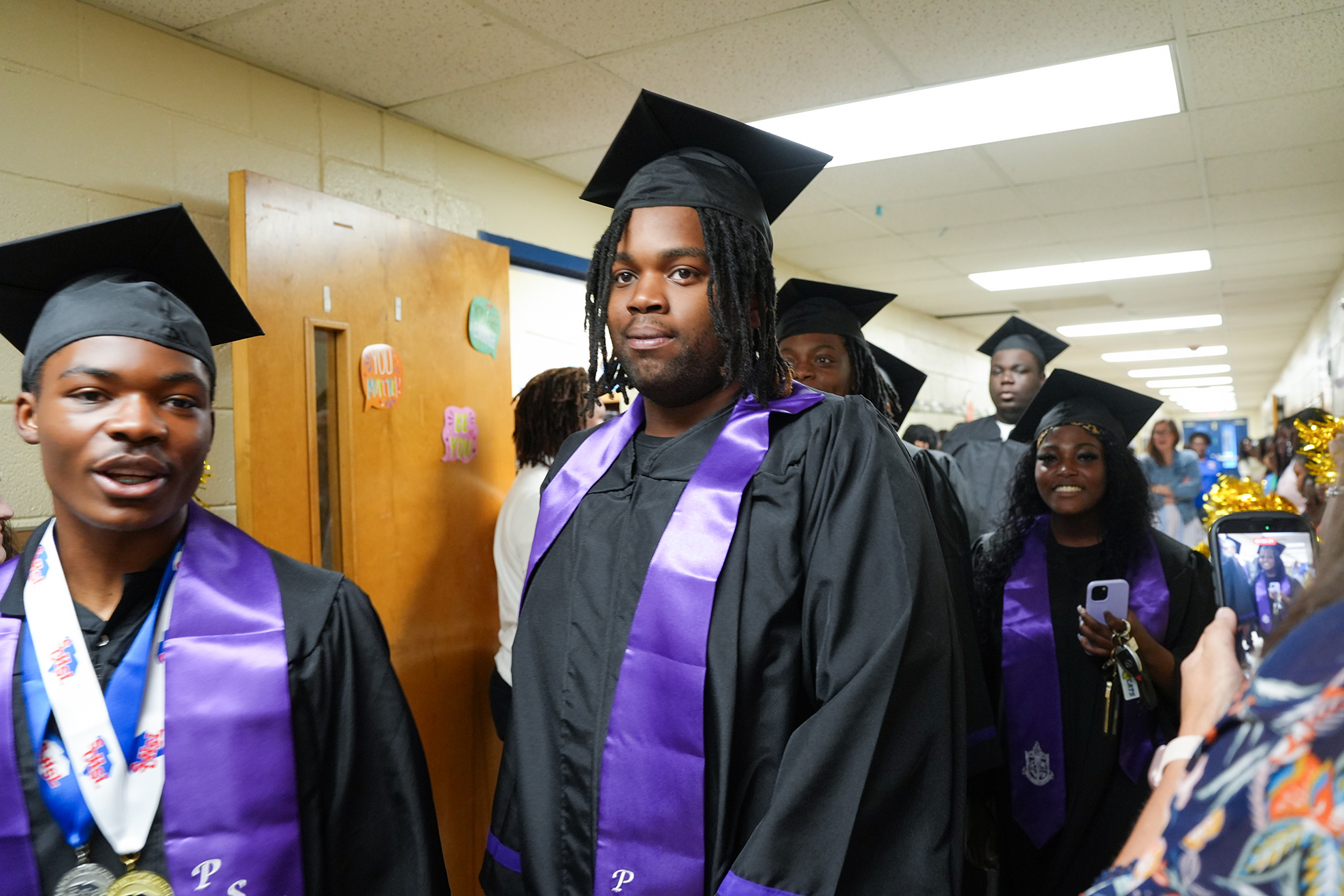 Graduates in caps and gowns walk down a hallway. One wears a medal and another holds a cell phone.