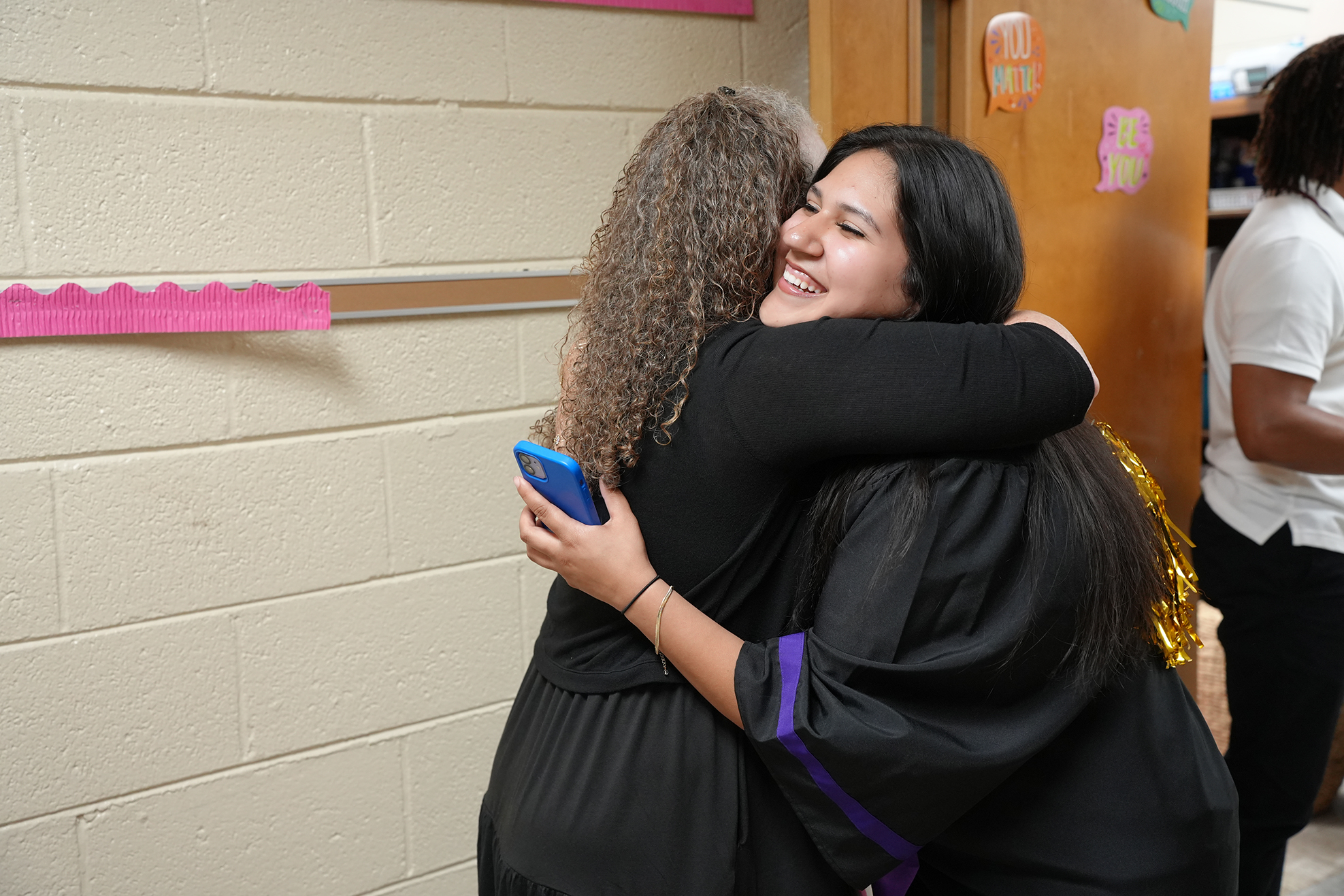 Two women embrace in a classroom, one holding a phone, smiling. White wall and door behind them.