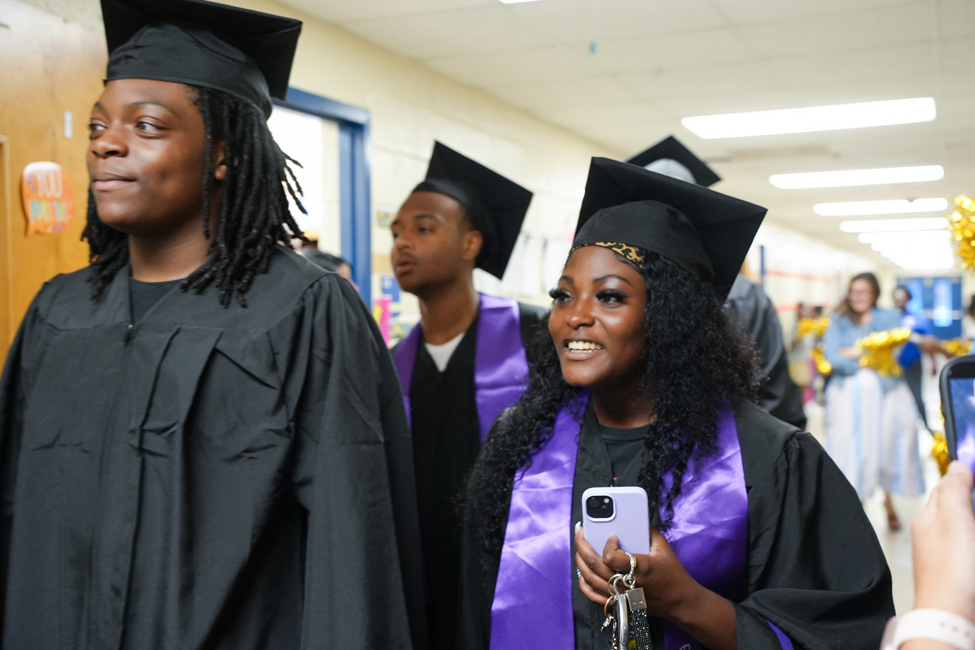 Three students in caps and gowns walk in a hallway. The girl in the center holds a phone.