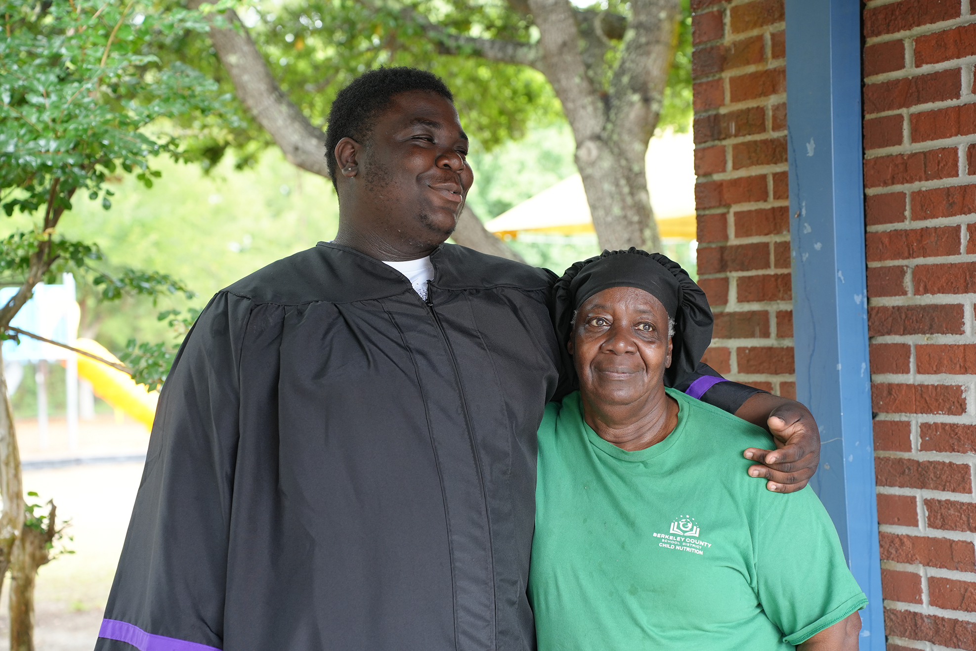 A man in a graduation gown poses next to a woman in a green shirt, standing against a brick wall with a tree behind them.
