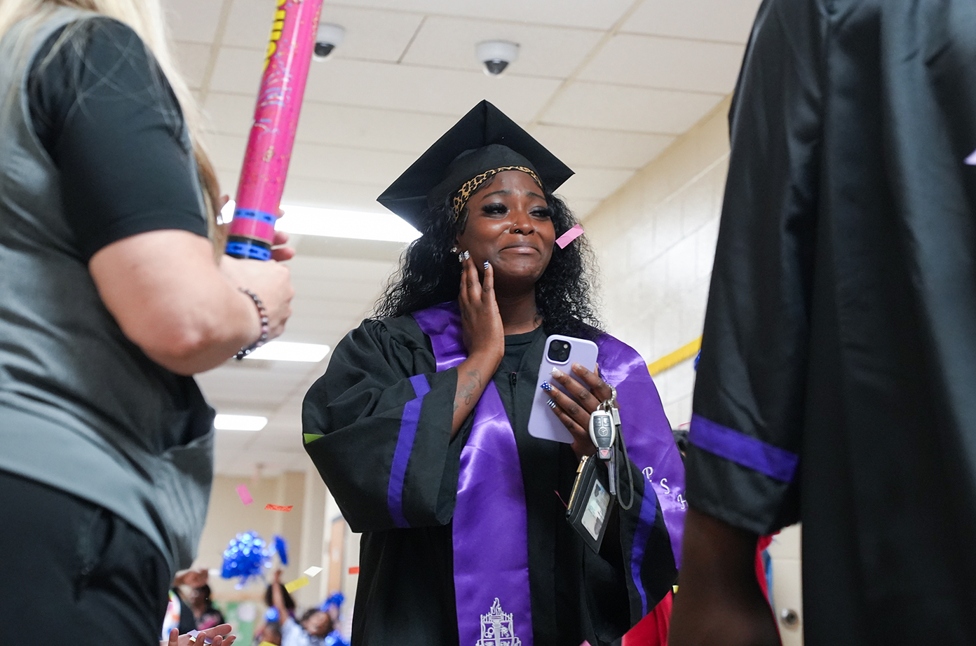 A person in a black graduation gown and cap holds a phone and speaks into it.