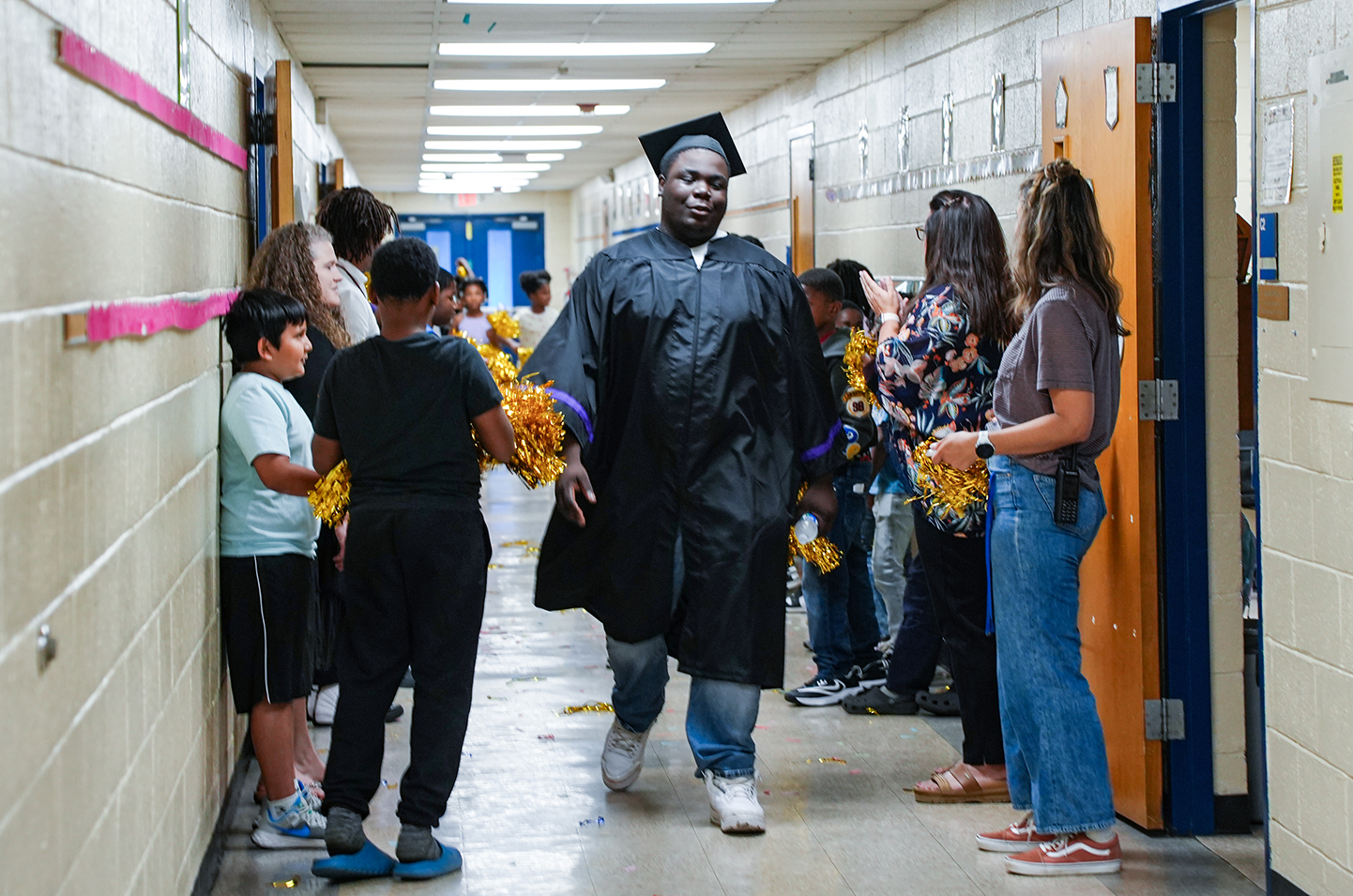 A person in a cap and gown walks in a hallway. Children and adults watch and cheer.