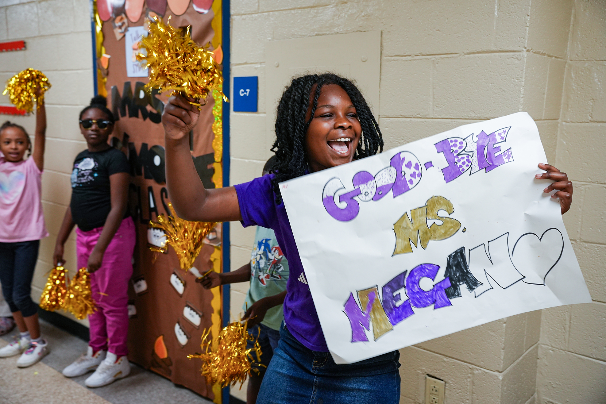 A girl holding a sign with purple letters saying "Good Vibes Only" and smiling. Behind her, two girls with pom-poms in front of a decorated door.