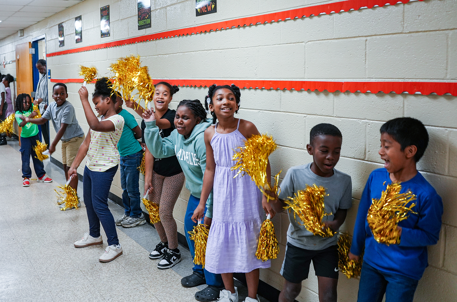 A group of children wearing school uniforms and holding yellow pompoms. They are standing against a white wall with red and orange trim.