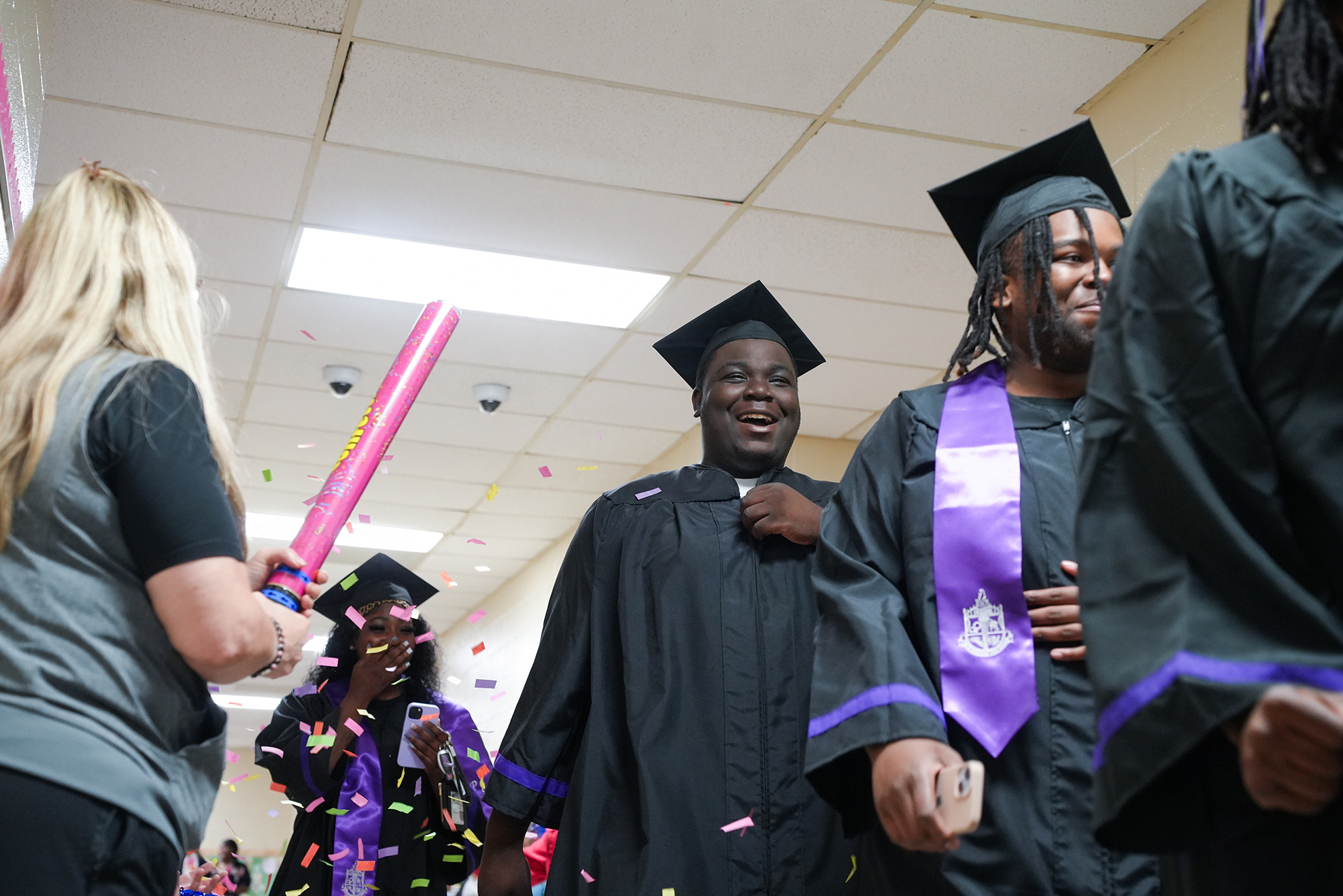 People in caps and gowns smile and walk. A woman in gray holds a pink object. Confetti falls from the ceiling.