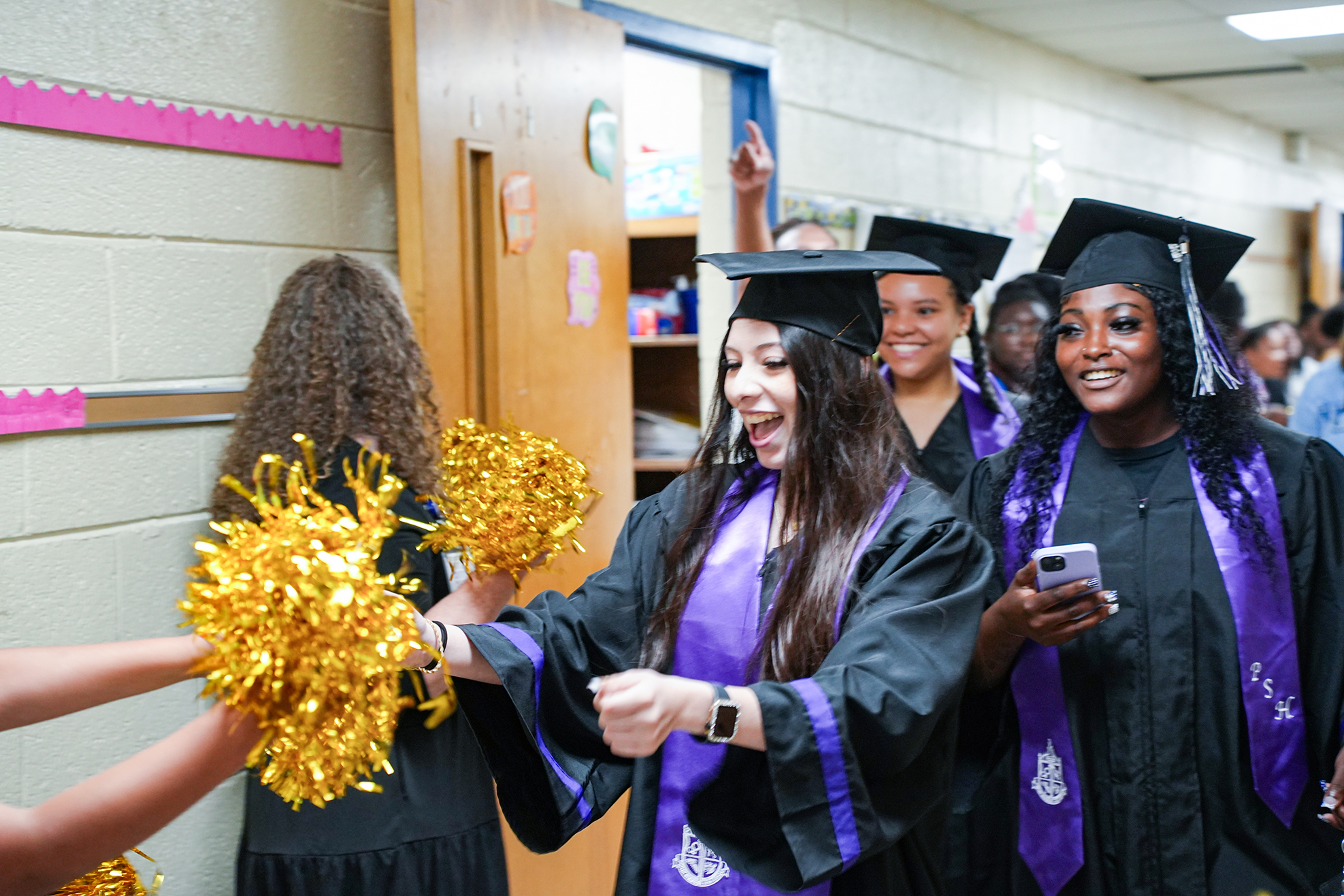 Graduates wearing caps and gowns walk down the hallway, one holding pom-poms, another on a phone.