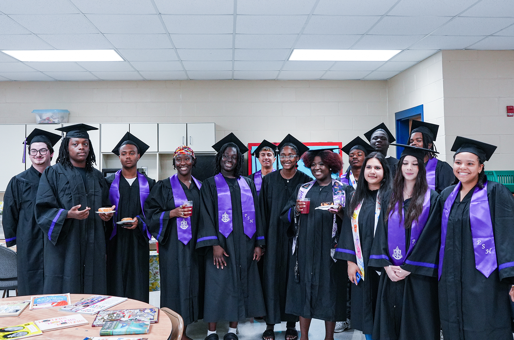 A group of individuals in graduation gowns and hats stand in a room. They hold cups and plates with food.