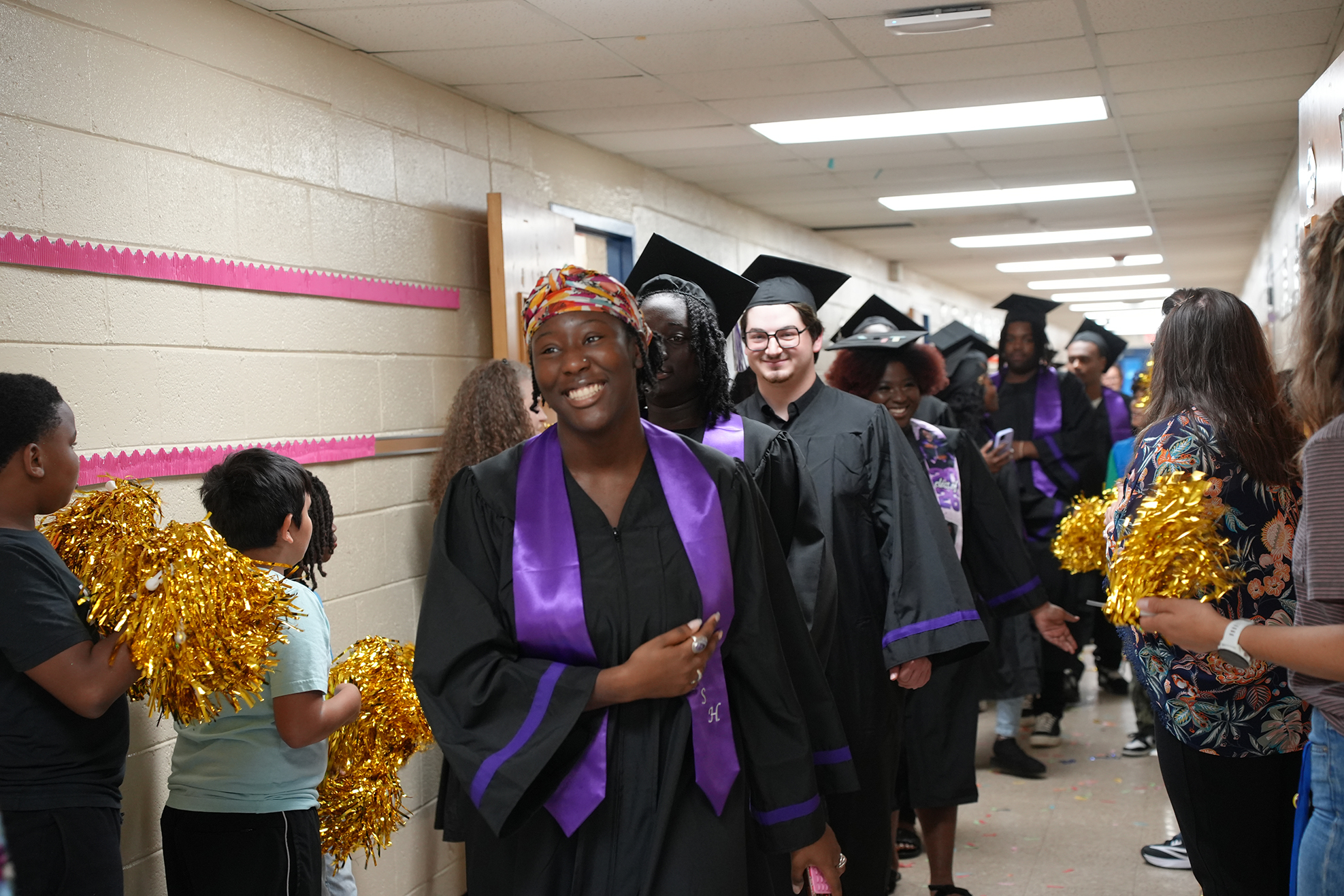 People in caps and gowns walk through a hallway, some holding pom-poms, while others look on.