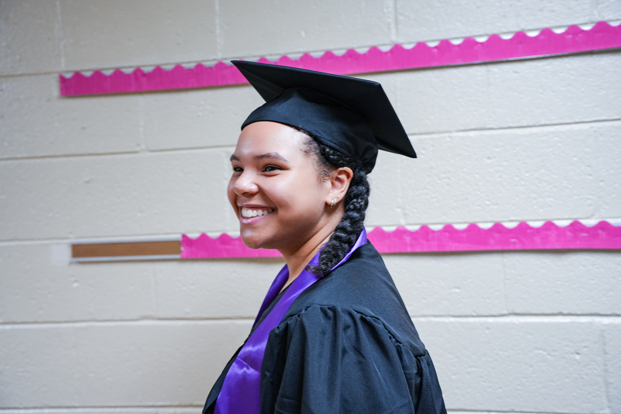 Person in a cap and gown smiling against a white wall with pink decorative borders.