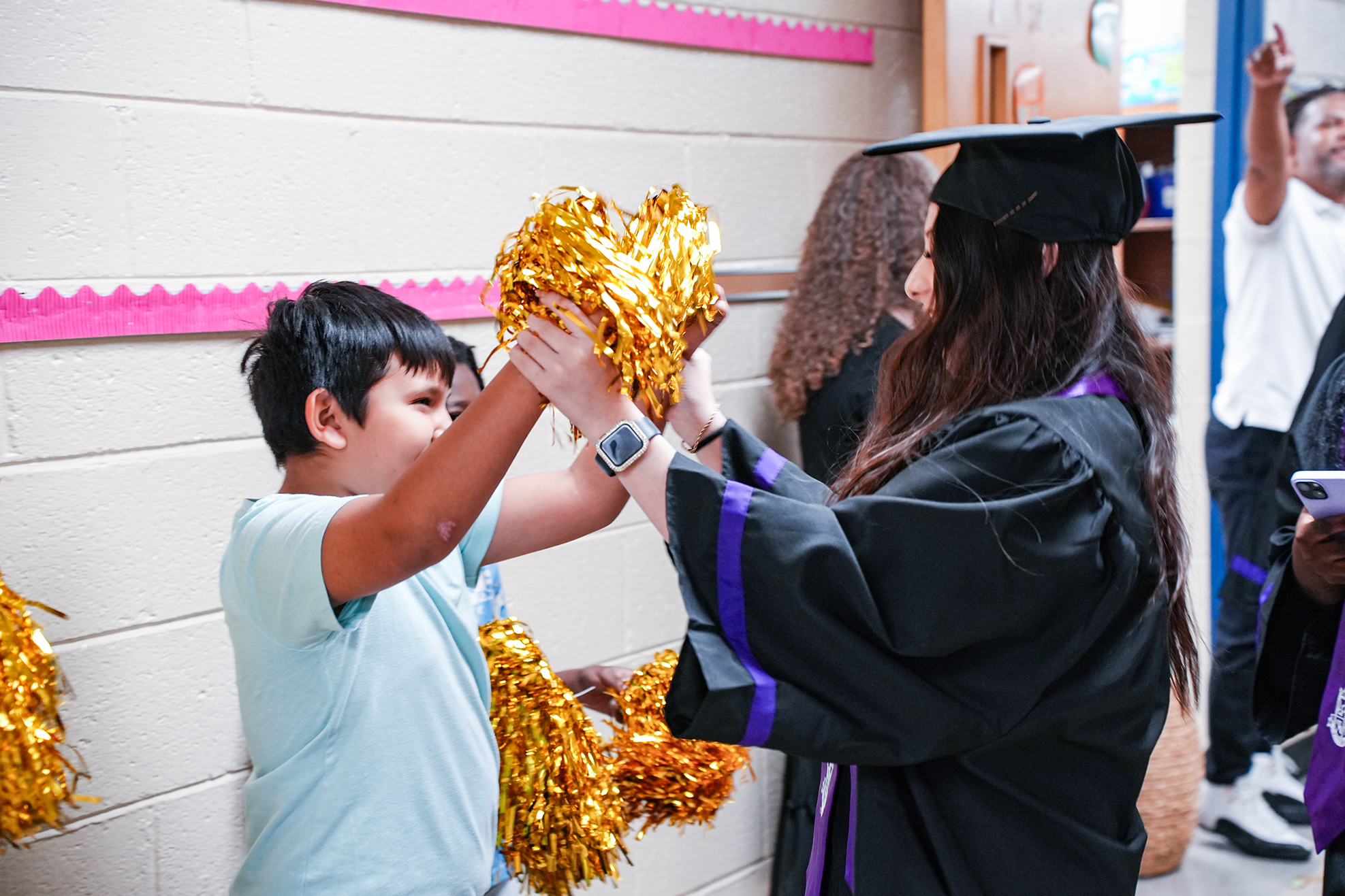 A young boy holding pom-poms helps a woman wearing a graduation gown and cap.