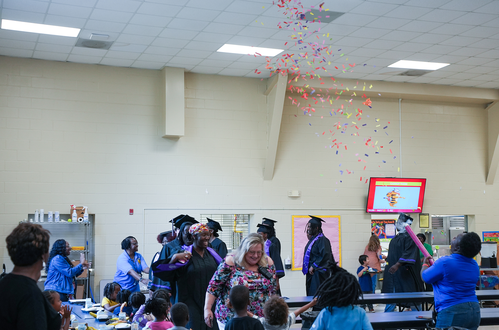 A room with a table and chairs, a screen, and people in graduation robes with confetti falling.