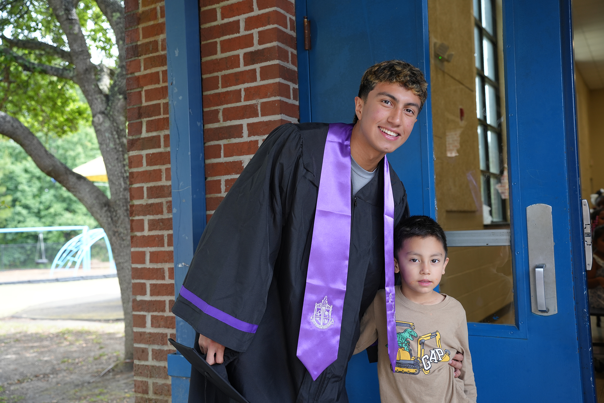 A boy in a graduation gown stands beside a younger boy in front of a blue door with a brick wall.