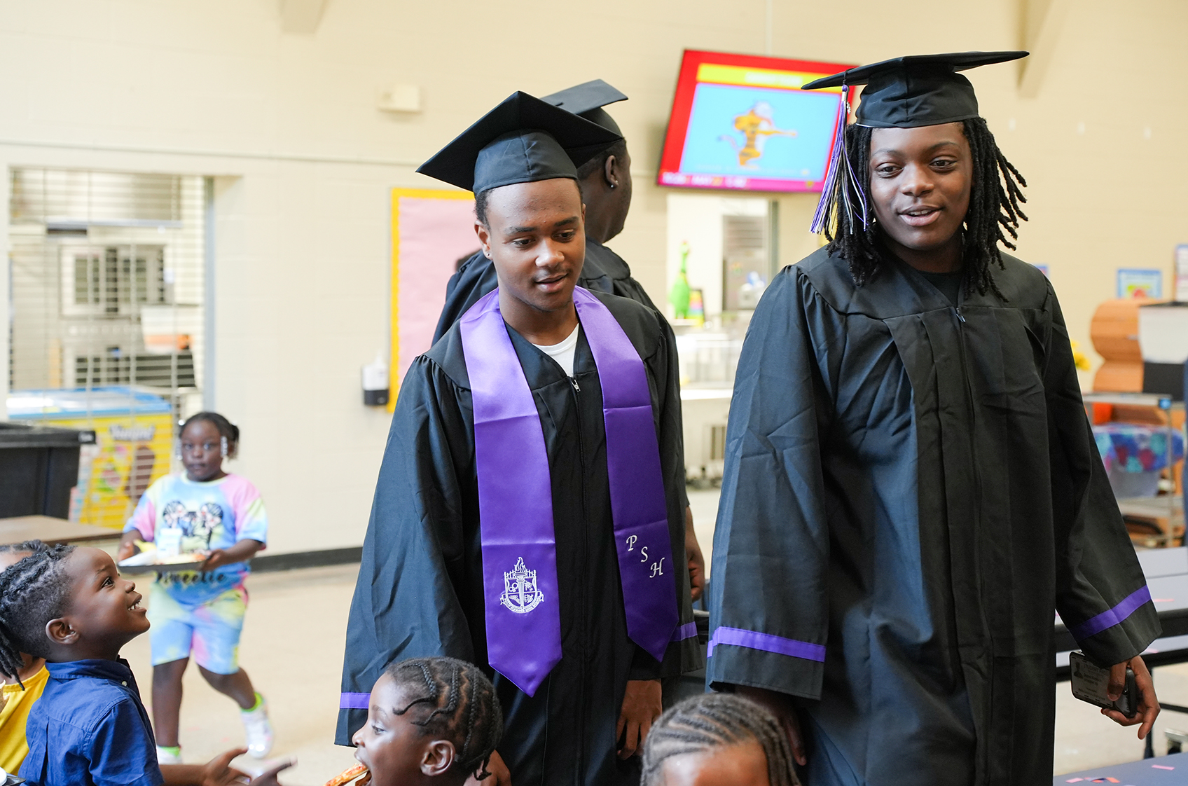 Graduates in academic regalia stand next to children. One holds food. A TV screen is in the background.