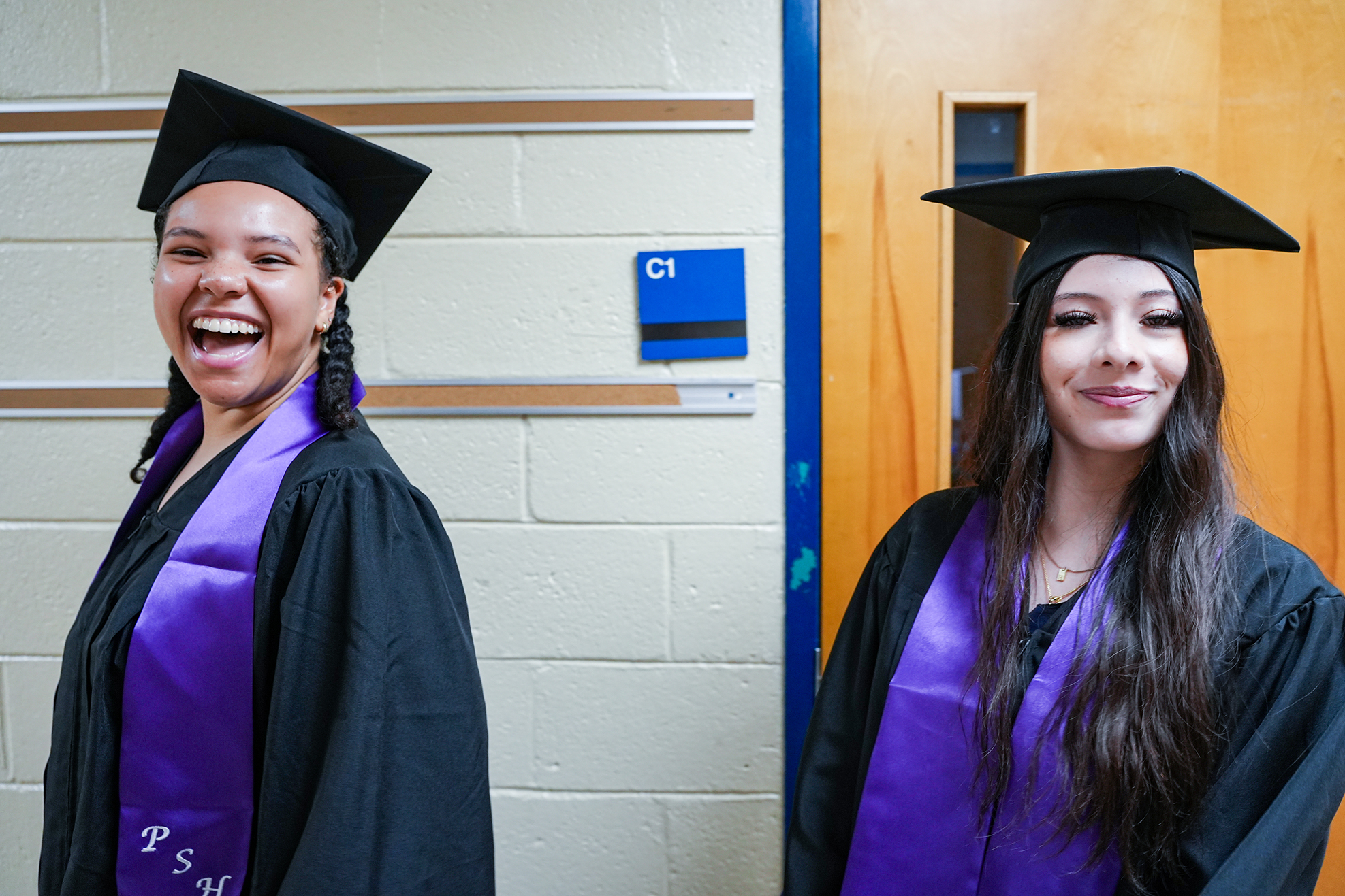 Two women in graduation robes and caps stand side by side, smiling. One has braided hair, and both wear purple stoles.