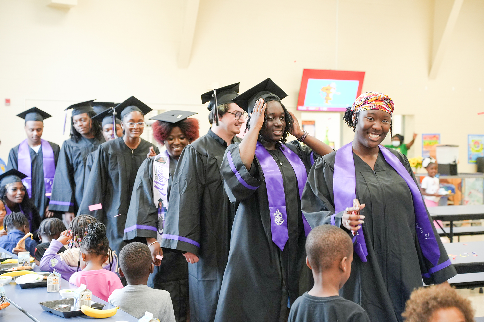 Graduates in caps and gowns stand in line, smiling, with children seated at tables in the background.