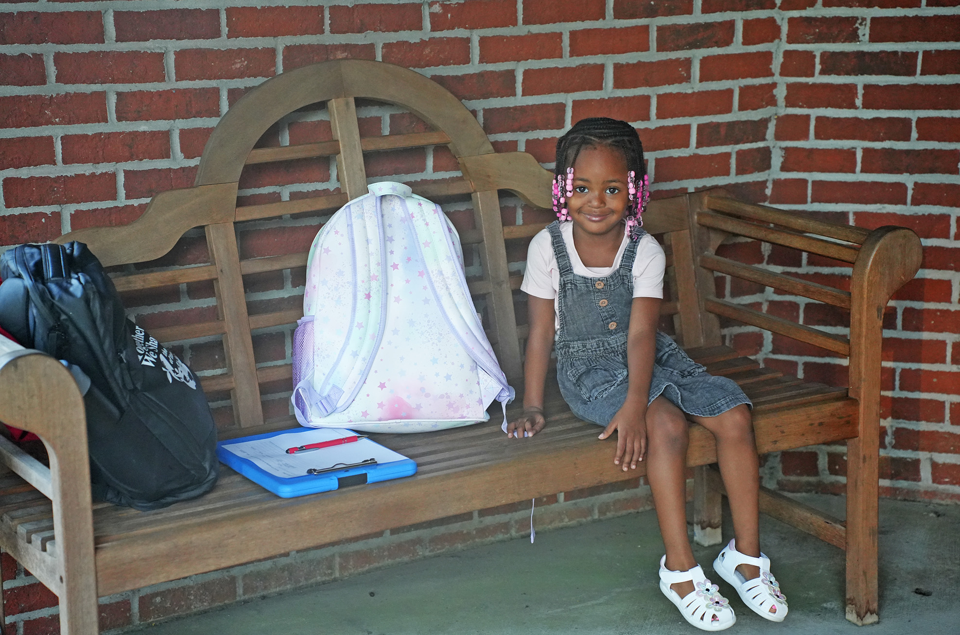 A young girl in a dress and sandals sits on a bench with a backpack and a folder.